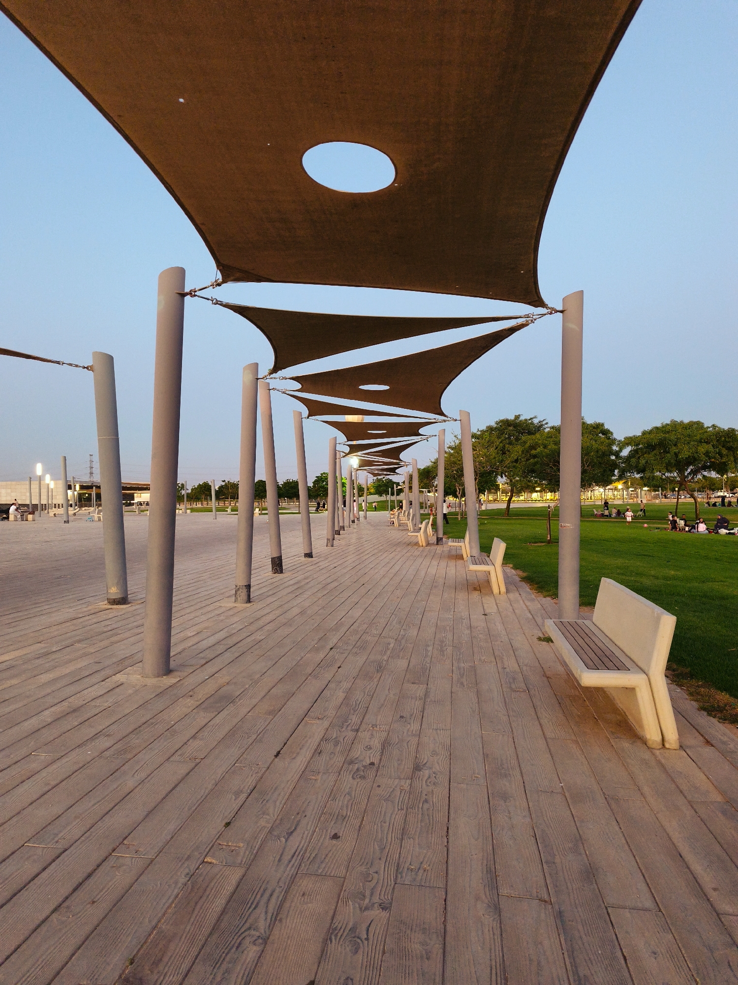 The image depicts an outdoor seating area with a wooden boardwalk and shade structures, located in Be'er Sheva, Israel. The boardwalk is paved with wooden planks and features several concrete benches with wooden slats. Overhead, a series of brown fabric shade sails are supported by tall, gray metal poles, creating a covered walkway. The sails have circular cutouts that allow some sunlight to pass through. To the right of the boardwalk is a lush green lawn where several people are seen sitting and relaxing. Beyond the lawn, there are trees and a building in the background. The sky is clear and light blue, indicating it is likely late afternoon or early evening. The overall scene suggests a public park or recreational area designed for people to relax and enjoy the outdoors.