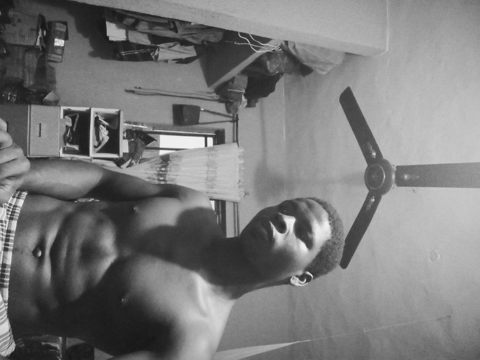 A black and white, waist-up, medium shot photo taken indoors in Nkpor, Nigeria shows a shirtless Black male looking directly at the camera. He has short, dark hair, and is wearing checkered shorts. The background of the image is dimly lit and cluttered, with a ceiling fan visible overhead. To the left, there is a shelf with clothes and bags, and to the right, a small cabinet with items on its shelves. The man's torso is well-defined and lit by a light source from his right, casting shadows on his left side. The overall impression is that of a casual self-portrait taken in a bedroom or storage area.