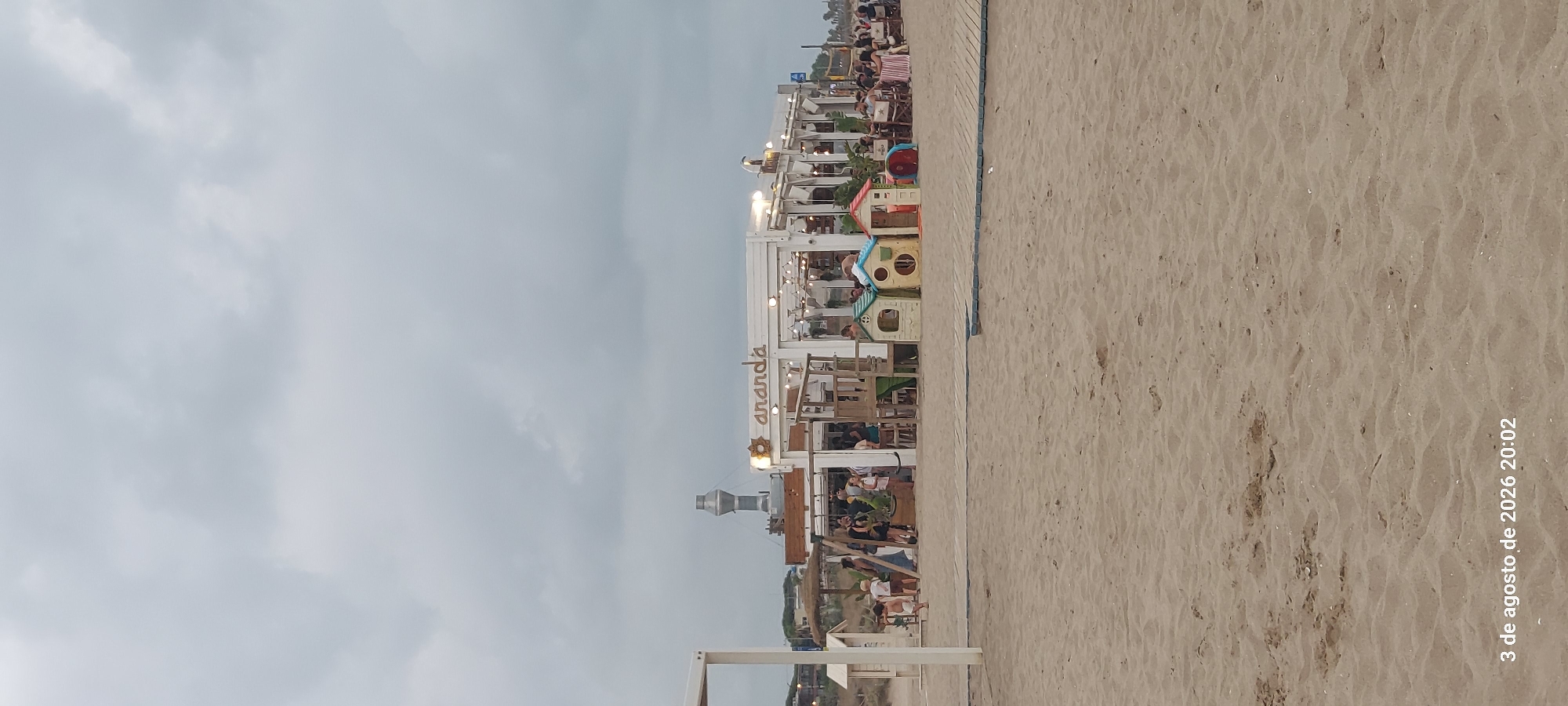 The image depicts a view of a beachside establishment with the name "ananda" visible. The establishment appears to be a restaurant or bar with outdoor seating. There are people seated at tables and standing around. In the foreground, there is a sandy beach with a wooden walkway. In the background, there is a tall structure that resembles a lighthouse or industrial chimney. The sky is overcast. The timestamp "3 de agosto de 2026 20:02" is visible in the bottom left corner, indicating the image was taken on August 3, 2026, at 8:02 PM. This location is consistent with the beach area of Castelldefels, Spain, known for its beachside businesses.