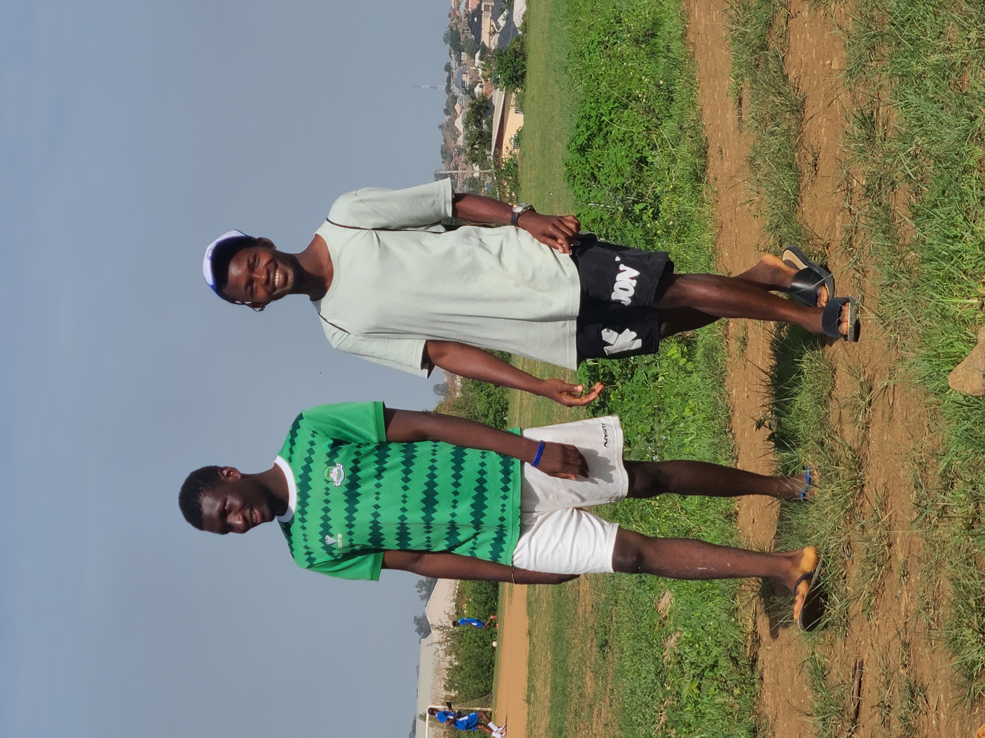 Two men are standing on a grassy and dirt field in a city or town. The man on the bottom is wearing a green and white patterned short-sleeved shirt and white shorts. The man on the top is wearing a light gray t-shirt and black shorts. Both are wearing sandals. In the background are buildings and vegetation. The sky is blue.