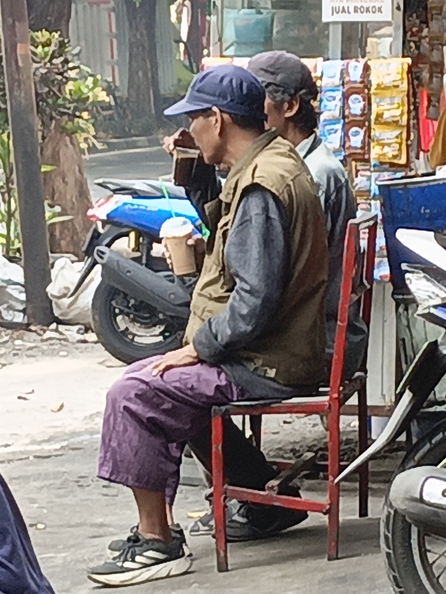 Two men are seated on a red chair on a street in East Jakarta, Indonesia. The man in the foreground, wearing a blue cap, brown vest, and purple pants, holds a cup of coffee. The man behind him, wearing a dark cap and a light-colored shirt, is partially visible. A blue motorcycle is parked behind them, and a rack of goods is visible in the background, suggesting a roadside stall. The ground is paved, and trees and foliage are present in the environment.