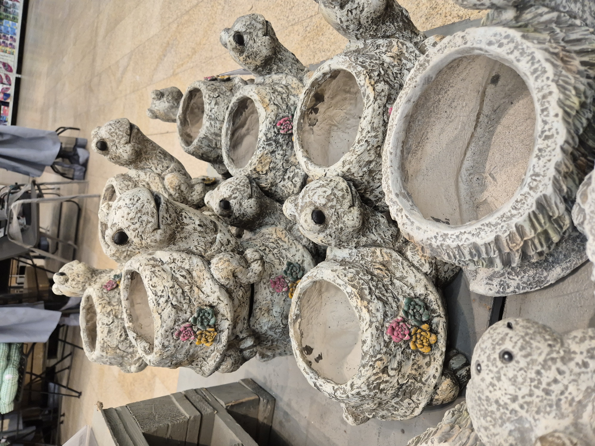 The image shows a collection of decorative turtle planters, likely indoors due to the presence of shopping carts and a neutral-colored wall. The planters are made of a textured, off-white material and feature turtle heads and limbs integrated into their design. Each planter has a circular opening, presumably for holding plants. Some planters are decorated with small, colorful flower appliques. The scene does not provide enough information to establish a location in Moscow, Russia.