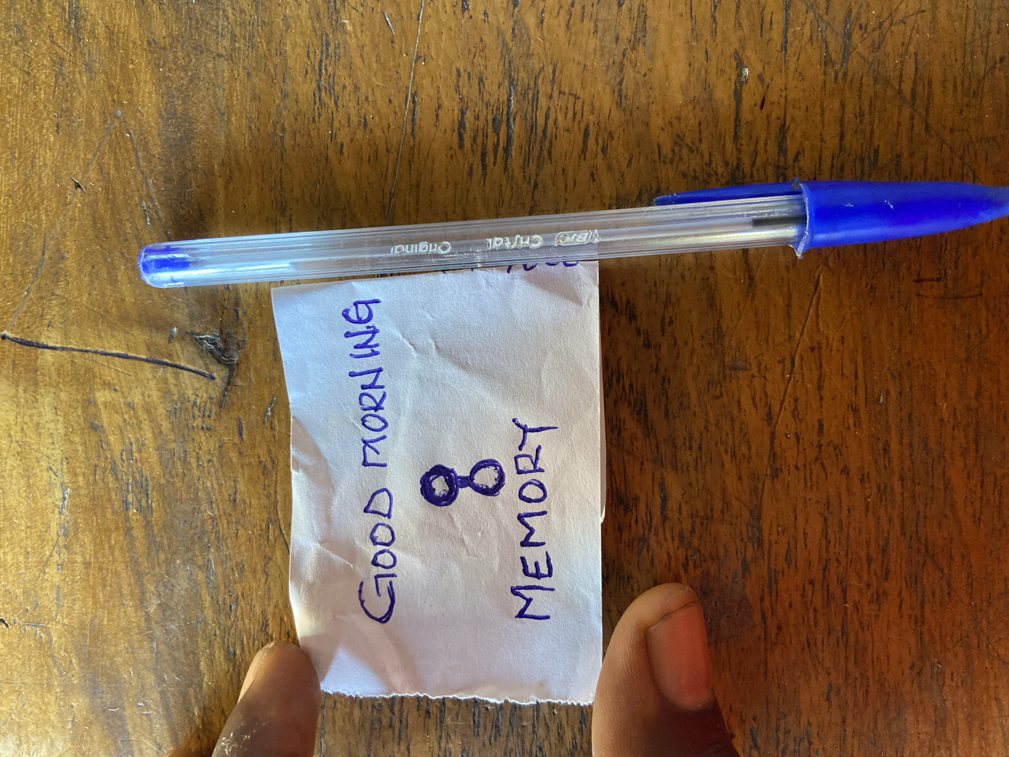 The image shows a close-up, overhead view of a piece of crumpled white paper with handwritten text in purple ink, placed on a wooden surface. The text reads "GOOD MORNING" on the top line, with a symbol that looks like two connected circles or an hourglass below it. The bottom line reads "MEMORY". A blue BIC Cristal Original pen is positioned vertically next to the paper. A person's thumb and index finger are visible on the left side of the paper, holding it down. The wooden surface appears to be a table with visible grain and some scratches. The overall lighting suggests an indoor or shaded outdoor environment. The location of Mararaba, Nigeria, is not visually identifiable from the image itself.