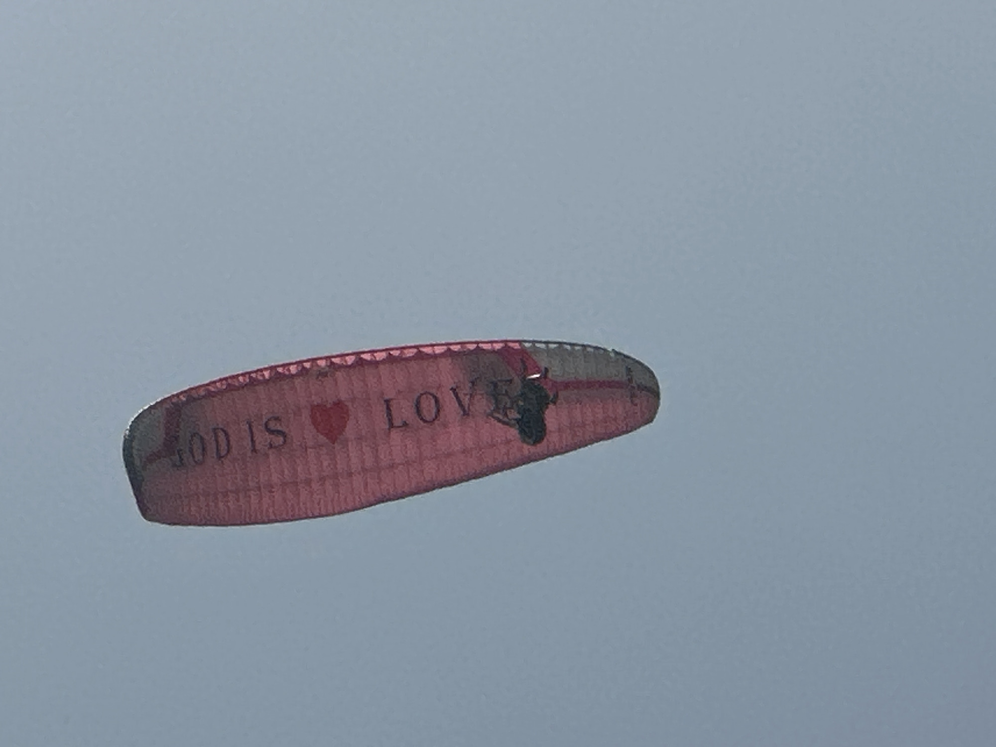 An extreme upward angle shot shows a pink paraglider with "GOD IS LOVE" printed in black on its fabric against a pale blue sky. The paraglider has a red heart symbol between the words "GOD IS" and "LOVE". A person is visible suspended beneath the paraglider, appearing to be in mid-flight. The paraglider is in the upper right portion of the frame. The image's context as being taken in Budva, Montenegro, is not visually apparent.