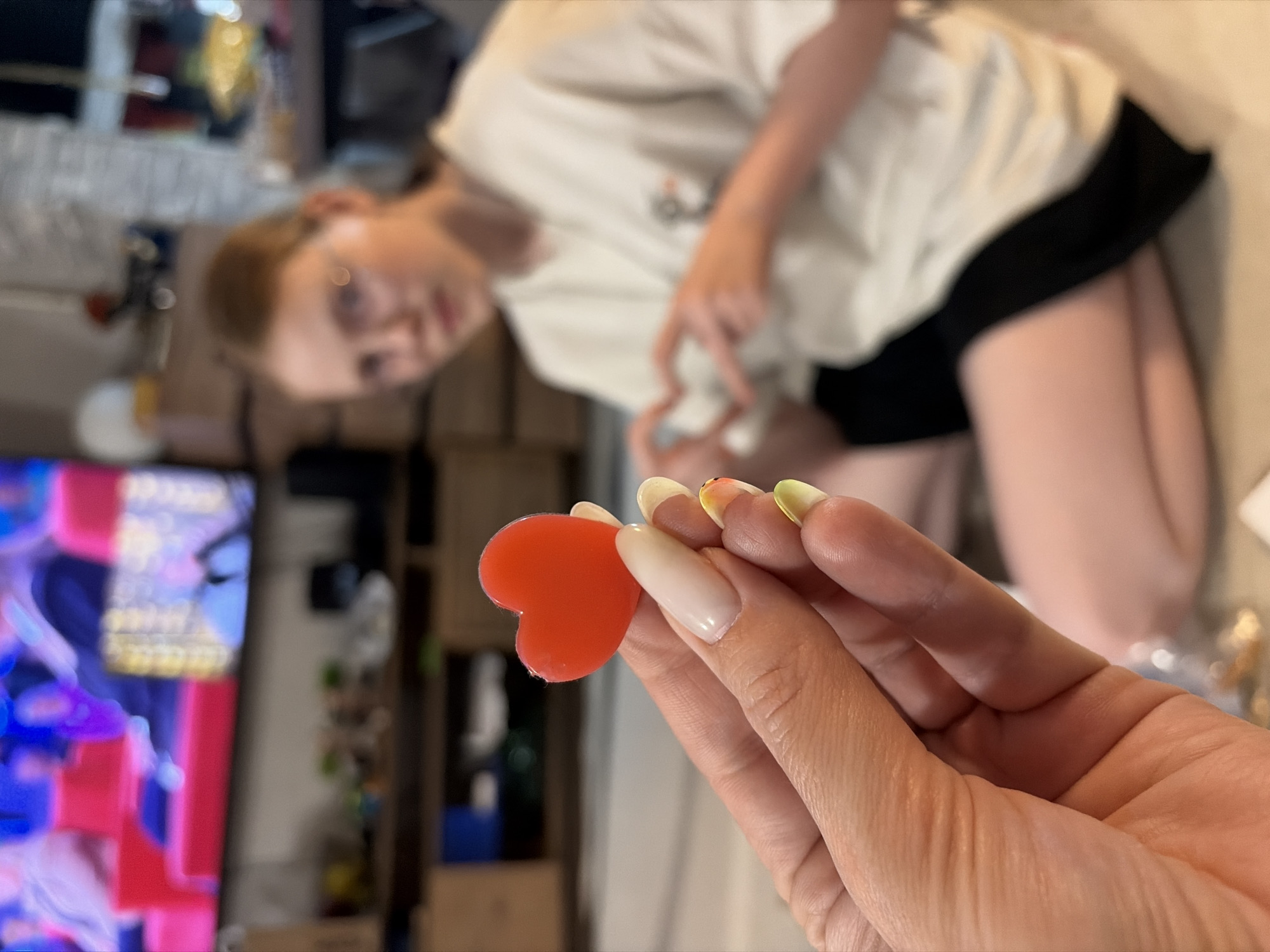 A first-person shot shows a hand holding a red heart-shaped gummy candy. The hand has a long, oval nail shape, with white, yellow, and green nail polish colors. Behind the hand, a blurry image shows a person wearing glasses, a white t-shirt, and black shorts, kneeling on the floor. A television screen displaying colorful images is also visible in the background. The scene appears to be inside a home. The location is Chaikovsky, Russia.