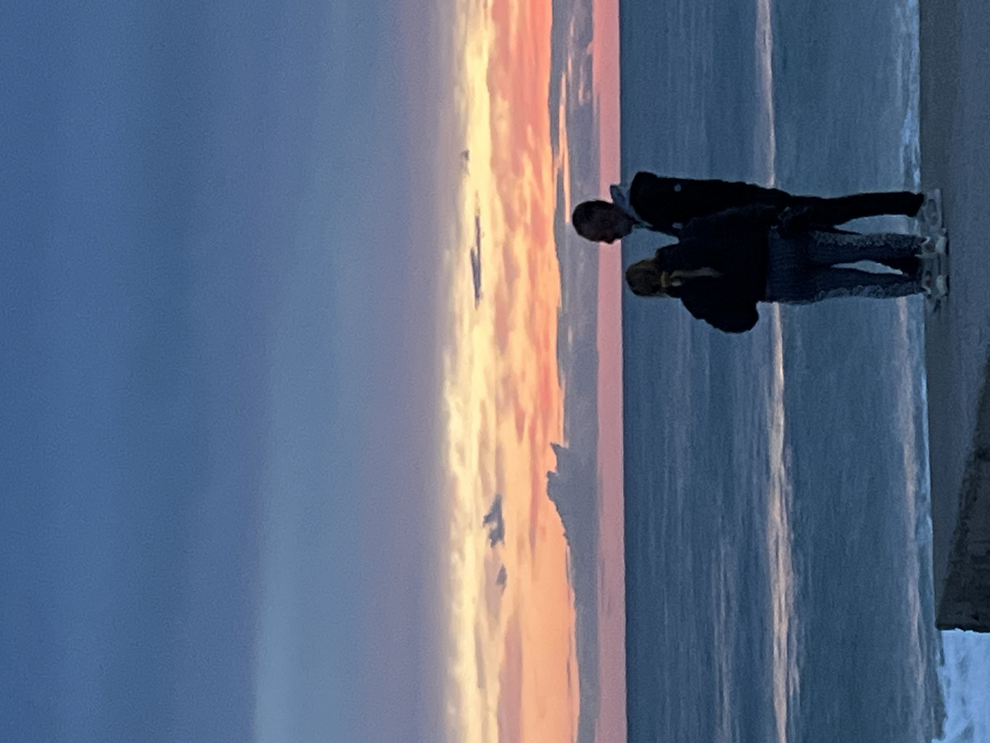 Two people, an adult and a child, stand side-by-side looking down at something. The adult is wearing dark pants and a jacket. The child has on a jacket and patterned pants. They are next to a body of water with ice or snow. The sunset sky has a blend of pink, orange, and yellow hues with clouds. A blue-toned sky is also visible. The location is Alanya, Turkey.