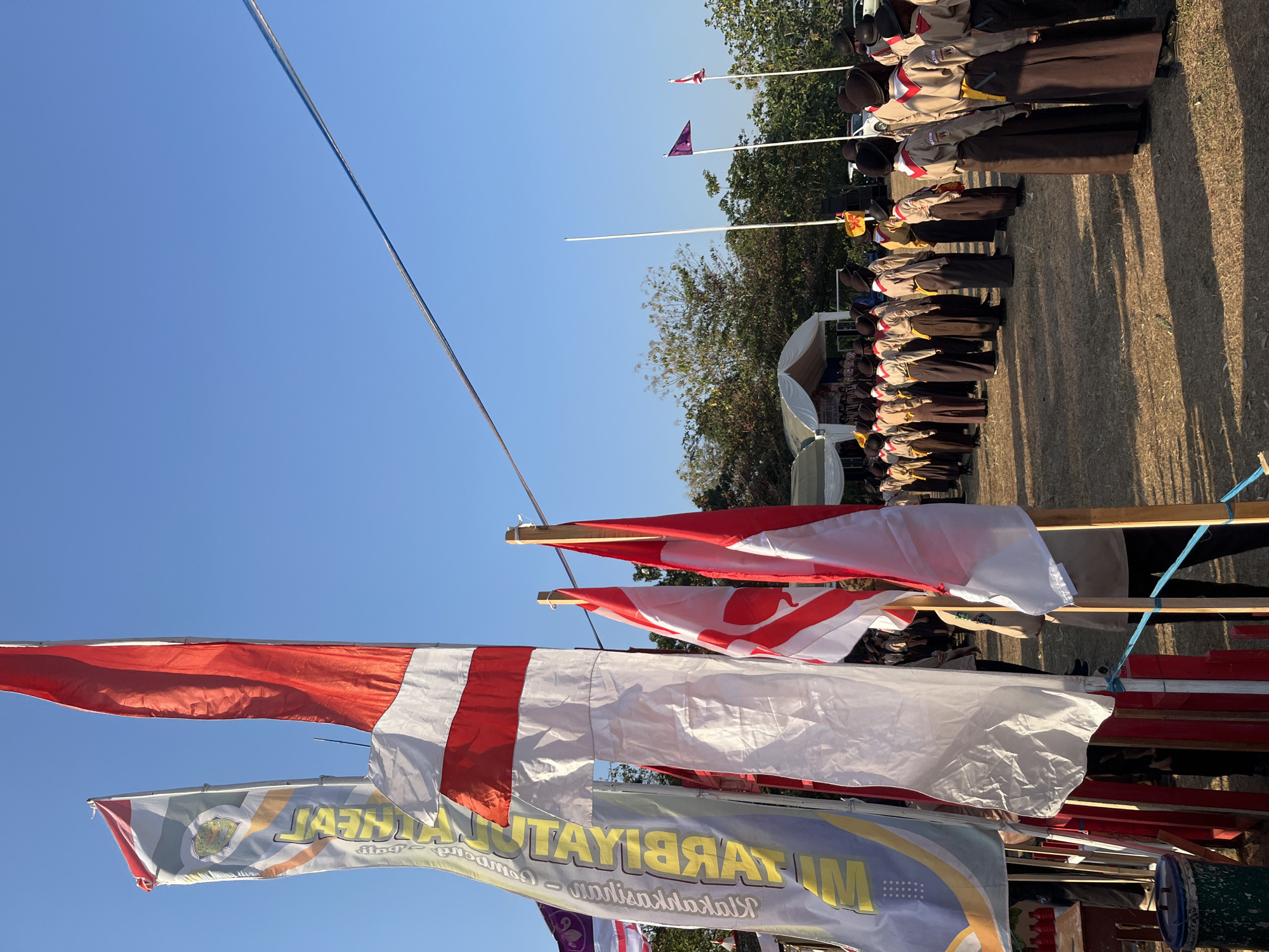The image captures an outdoor event, likely a ceremony or gathering, under a clear blue sky. In the foreground, several flags are prominently displayed. One is a large red and white striped flag, appearing to be the Indonesian flag, draped and secured by wooden poles. To its left is another flag, predominantly white with red accents, and further left, a tall banner with yellow text on a grey background is visible. These flags are arranged in a line, suggesting a formal setup.
In the middle ground, a group of individuals, appearing to be scouts based on their uniforms, stand in formation on dry, grassy ground. They are dressed in light brown tops and dark brown skirts or trousers, with some wearing dark hats. Behind them, a white tent structure is set up, possibly a stage or pavilion, with more people gathered underneath. Trees and foliage are visible in the background, with a few more flags on poles further in the distance.
The scene suggests a sunny day, with the light casting shadows on the ground. The presence of the Indonesian flag and the scout-like uniforms points towards a possible national or community event in Indonesia. The location is identified as Gembong, Indonesia.