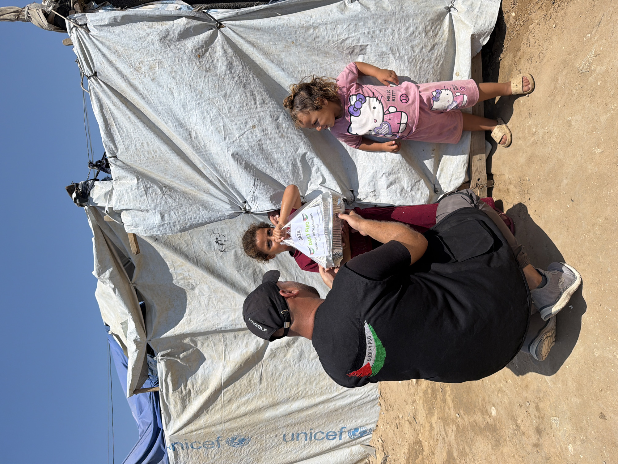 In Jabalia, Palestinian Territories, a man wearing a black vest with a Palestinian flag emblem and the text "DUDLEY PSG" kneels to hand a food container to a young girl. The girl, with curly hair, wears a maroon dress and holds the container, which has text including "GAZA DAILY FEED" and "SEPTEMBER 2020". Behind her, another young girl in a pink Hello Kitty outfit stands to the right. They are positioned in front of a makeshift shelter constructed from white and blue tarps. The ground is dusty earth. A UNICEF logo is visible on the white tarp in the background. The sky is clear and blue.