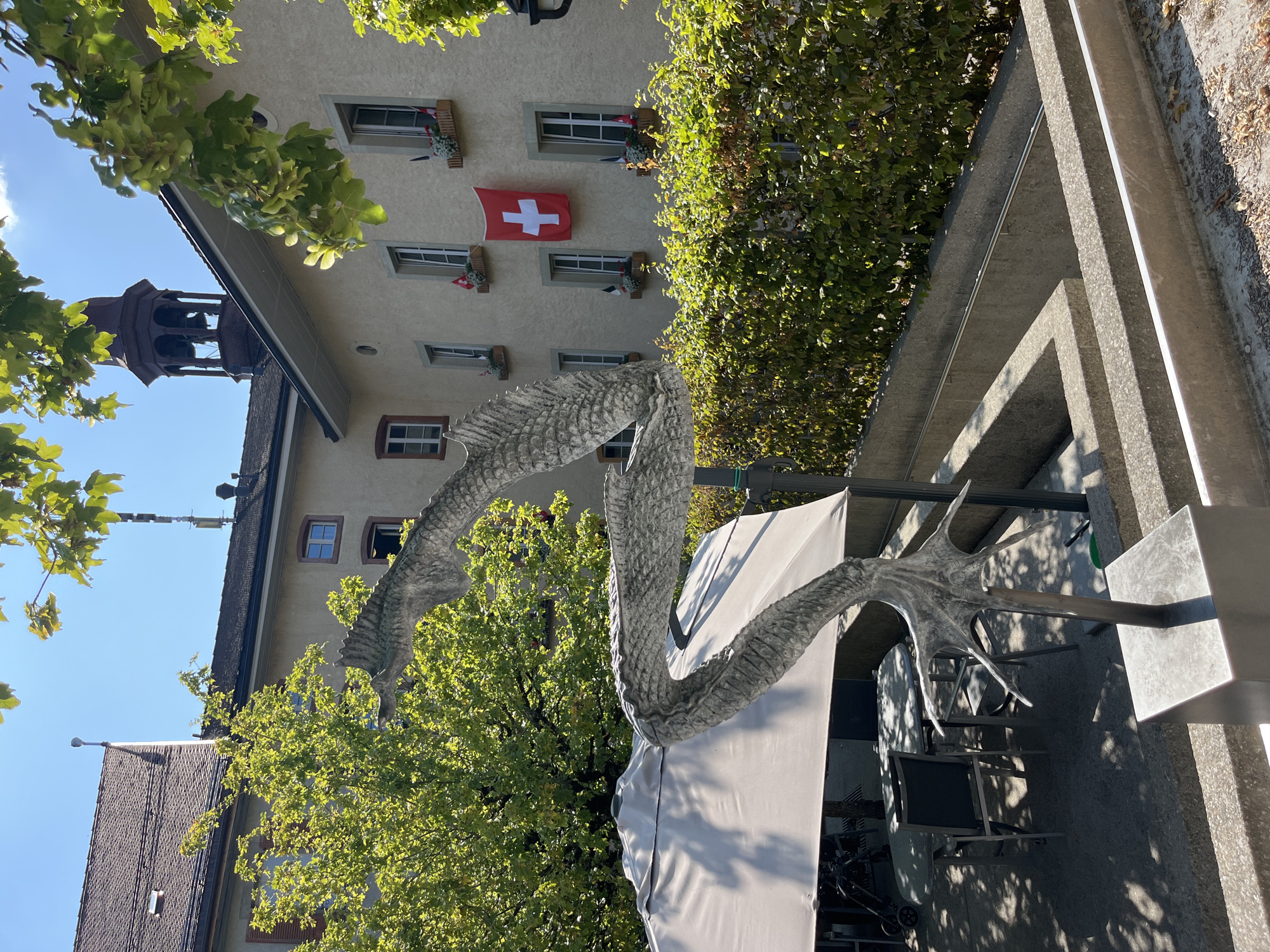The image depicts an outdoor scene in front of a building, possibly in Epagny, Switzerland, given the Swiss flag prominently displayed. In the foreground, a sculpture of a stylized dragon-like creature, made of grey metal, is positioned on a concrete base. The sculpture is partially obscured by the branches of a tree in the upper right corner.
Behind the sculpture, to the left, is an outdoor seating area with tables and chairs under a large, light-colored umbrella. The ground surface is paved with concrete. Further back, a dense green hedge lines the scene.
The building behind the hedge features a beige facade with several windows. Some windows have flower boxes filled with red and white flowers, and small flags. A large Swiss flag, a white cross on a red background, is hung on the wall between two windows. The building has a dark tiled roof. A belfry with a dark-colored dome and a bell is visible on the rooftop. The sky is clear and blue. The overall impression is one of a sunny day in an urban or semi-urban environment.