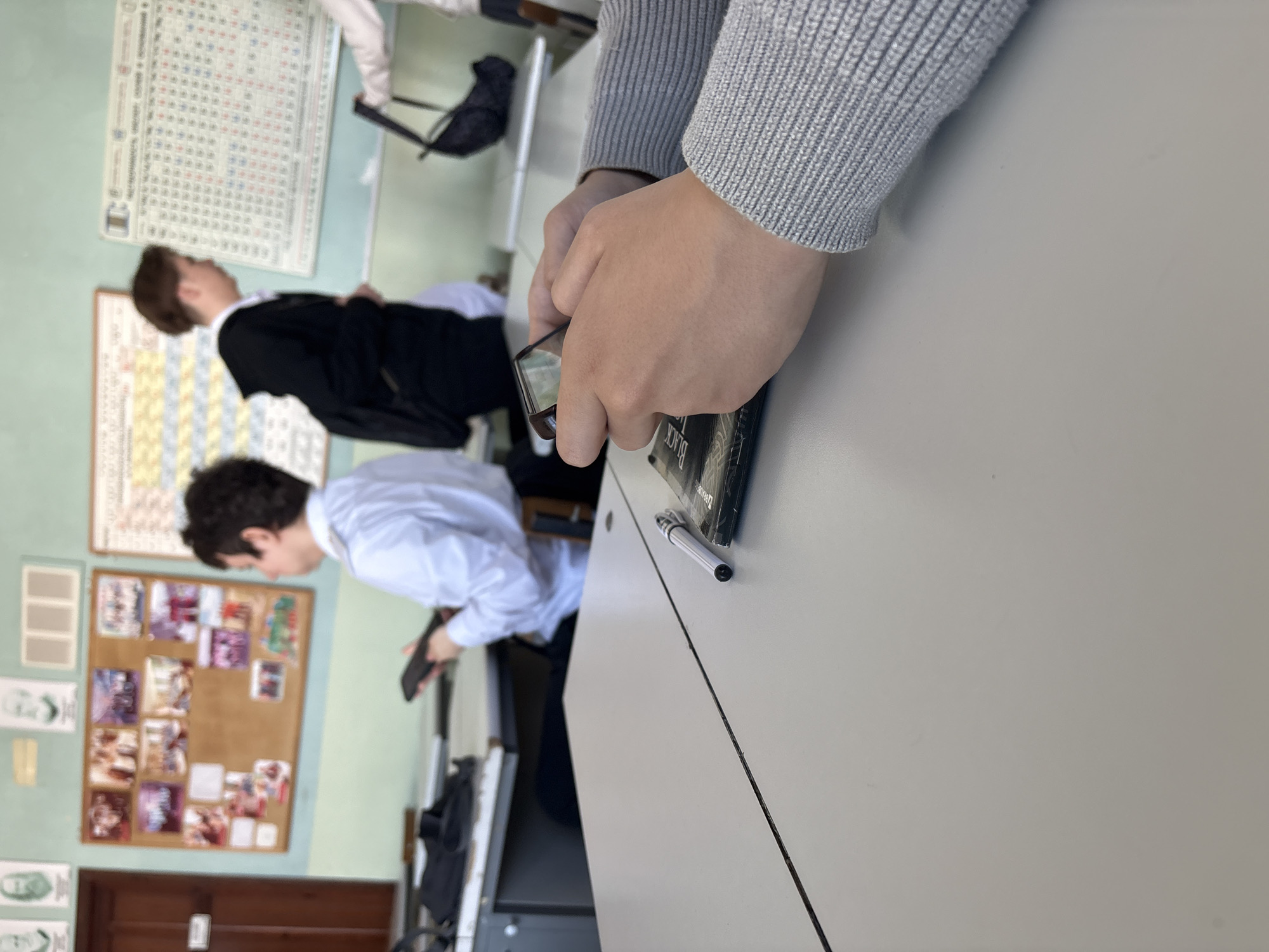 The image, taken from a slightly high angle, depicts an indoor classroom or educational setting. The scene is oriented vertically, with light green walls and light grey desks.
In the immediate foreground, a person's hands and forearms are visible, clad in a grey knitted long-sleeved top. Their hands are clasped around a black, rectangular object, possibly a smartphone or small book, on which the word "BLACK" is visible in white text. A white pen with a black tip lies on the light grey desk surface next to this object.
In the mid-ground, to the left, two male students are visible. One, wearing a black suit jacket and a white shirt, is looking towards the right side of the frame. Below him, another student in a light blue long-sleeved shirt is bending over, looking downwards towards the left. Black bags are partially visible beneath the desks.
The background features several items affixed to the light green wall. A large periodic table of elements poster dominates the upper left, with the title "Периодическая система элементов Д. И. Менделеева" (Periodic system of elements D. I. Mendeleev) faintly visible, indicating a Russian-speaking context. Below it, a smaller, blurred chart is present. A cork bulletin board adorned with numerous small photographs or pictures is prominent on the lower left wall. Below the bulletin board, a section of a wooden door is visible. The overall setting suggests an active learning environment, possibly during a break or a casual moment in class.
