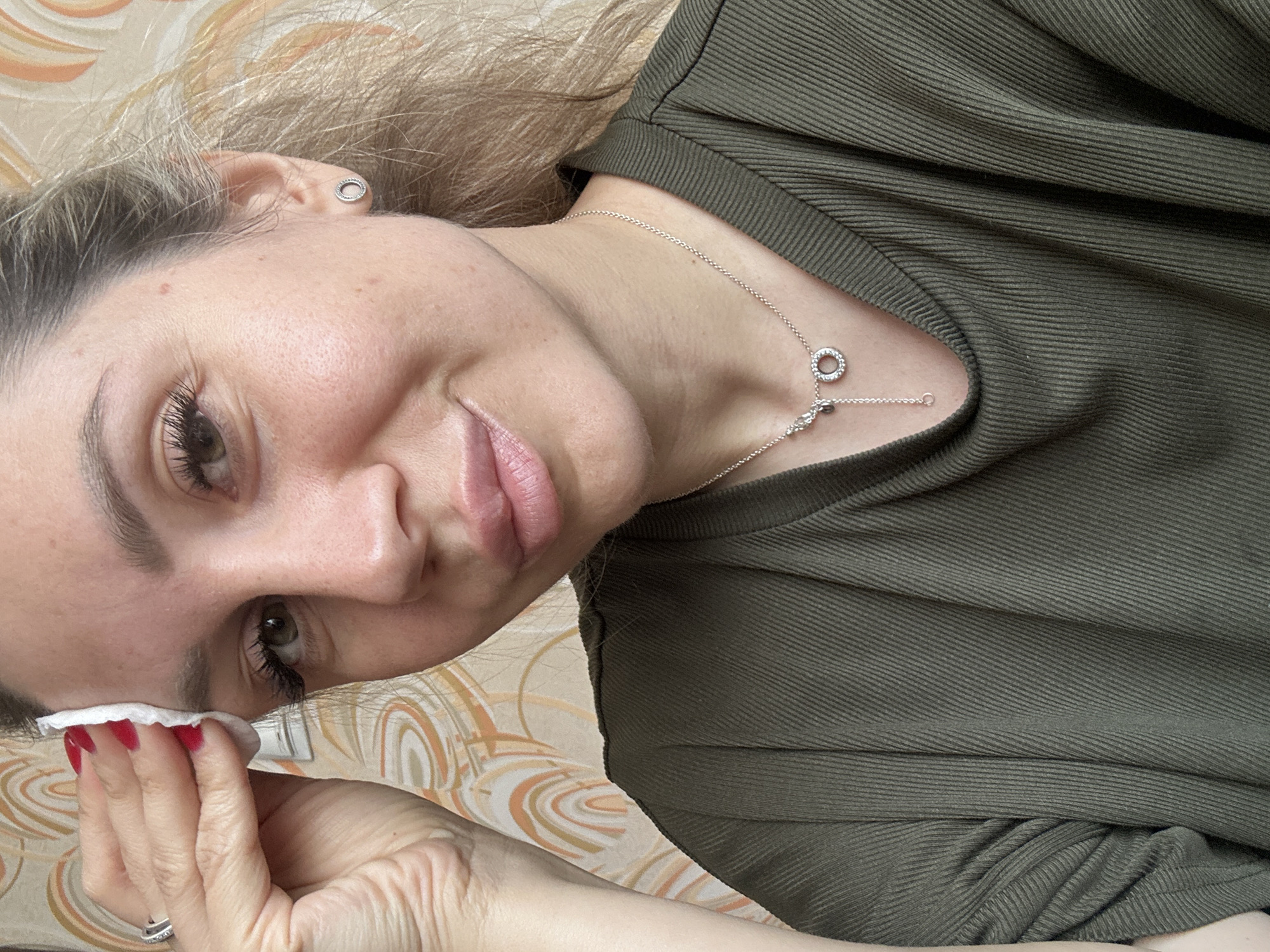 A woman with blonde hair tied back in a ponytail and wearing a green, ribbed t-shirt is holding a white cotton pad to her forehead. She has on a silver necklace with a circular pendant. She is also wearing a silver ring on her left index finger and a small silver stud earring in her right ear. The background is a patterned wallpaper. The image appears to be taken indoors. There is no information in the image to indicate a location in Novoaltaysk, Russia.