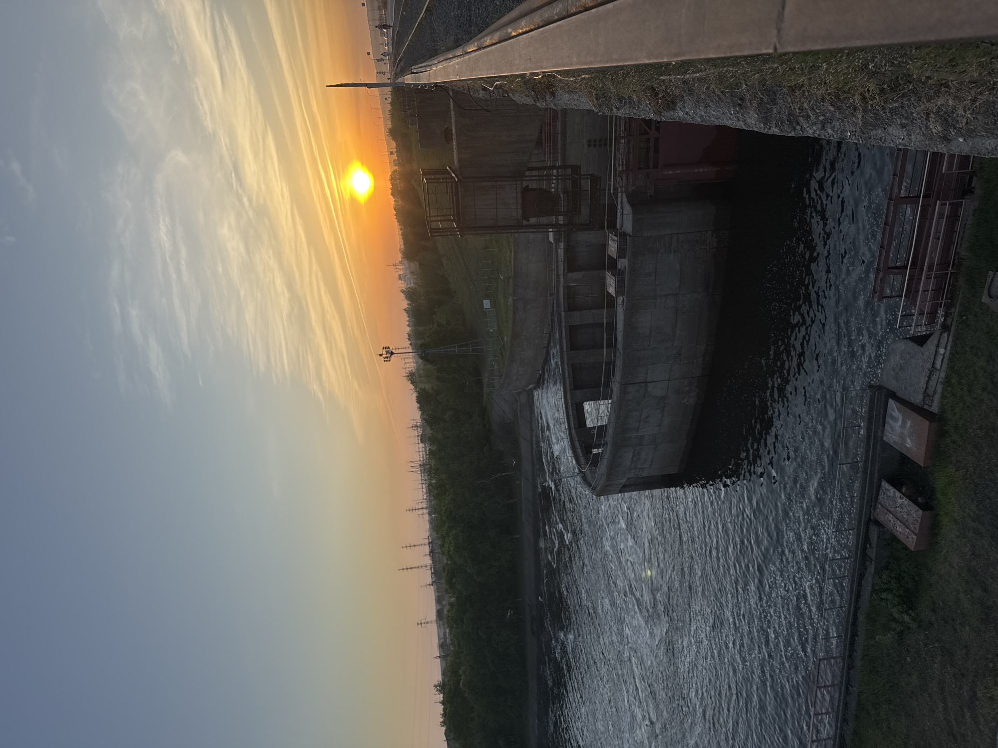 The image shows a canal in Chaikovsky, Russia at sunset. A concrete structure with a metal cage-like component extends into the water. Water is visible on the right side of the structure. Power lines and trees are visible in the distance. The sky is orange and blue with long, striated clouds near the setting sun. A metal railing is partially visible in the upper right of the frame.