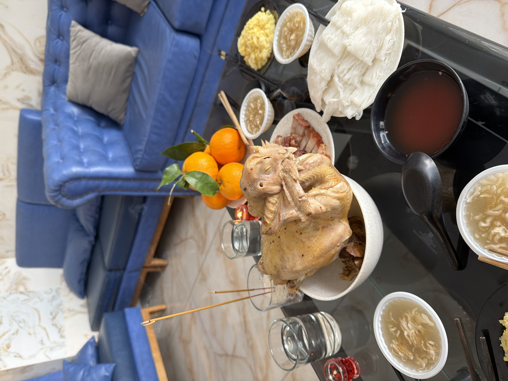 A spread of food and offerings is laid out on a black glass table, possibly for a ceremony or celebration. The scene is set indoors, with a backdrop of blue leather sofas and patterned wallpaper. In the foreground, a whole cooked chicken is presented in a white bowl, with its head slightly tilted. Surrounding the chicken are various dishes: a plate of sliced roasted meat, two small bowls of a clear, soupy dish with shredded ingredients, a plate of yellow scrambled eggs, and a pile of white, thin noodles. There are also small glasses of water, a red votive candle in a glass holder, and burning incense sticks in a small glass filled with what appears to be rice. Several oranges with green leaves are clustered in the background, near the sofas. The overall lighting is bright, suggesting daytime. The location is identified as Nha Trang Ward, Vietnam.