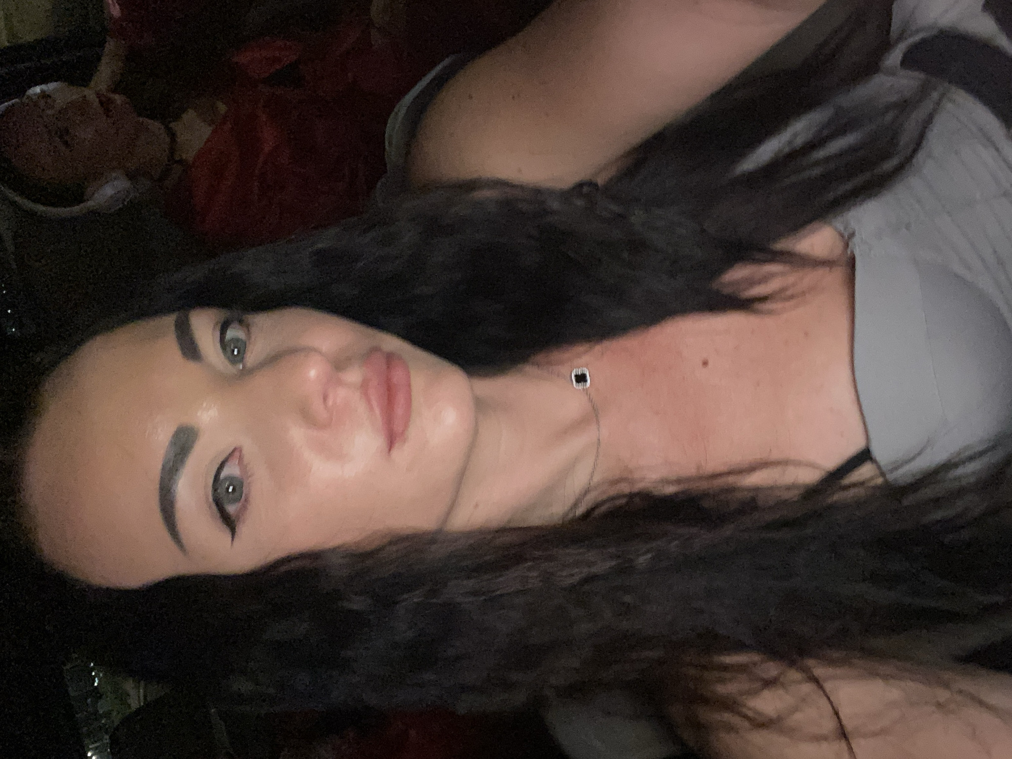In Sharm El Sheikh, Egypt, a woman with dark hair and noticeable makeup poses for a selfie. She is wearing a light gray top and a necklace with a black pendant. In the background, another person with headphones on is visible. The lighting suggests an indoor or nighttime setting.