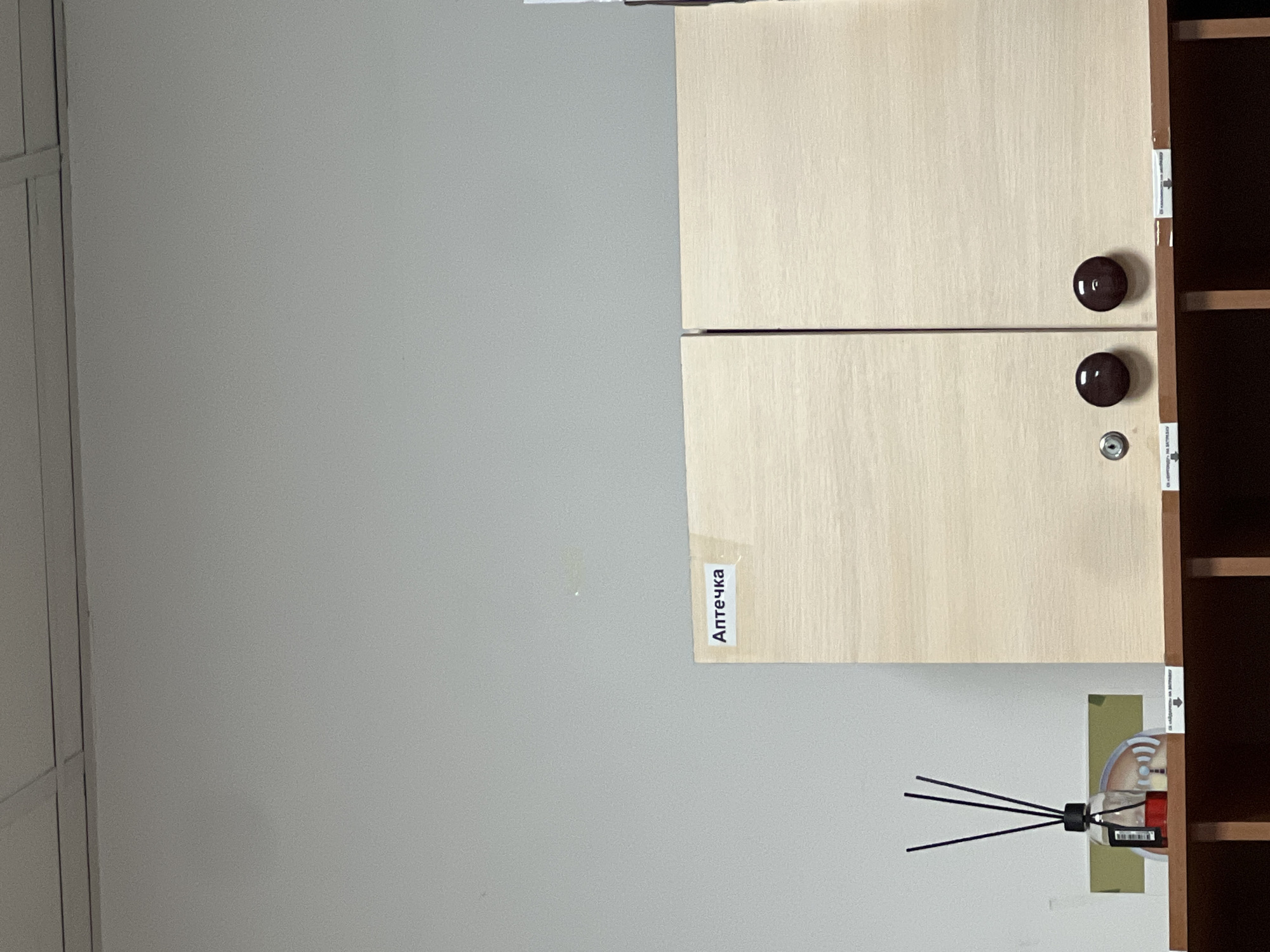 In the city of Kadamjay, Kyrgyzstan, a close-up, eye-level shot captures a segment of a room. The focus is on a light-colored wooden cabinet with two doors. A white label with Cyrillic text, "Аптечка" (meaning "First Aid Kit"), is affixed to the left door. Each door has a dark brown, circular knob for opening. A small, circular lock mechanism is visible to the left of the knobs on the left door. Below the cabinet, a wooden shelf is visible, supporting a glass bottle containing a red liquid and black reeds, indicative of a diffuser. The background consists of a light gray, textured wall, and a portion of a ceiling with visible tiles.
