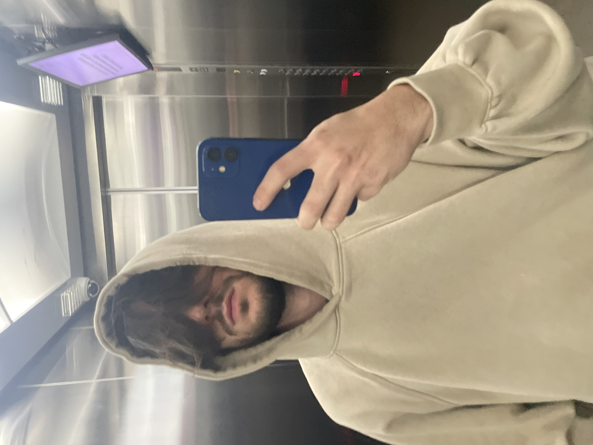 A person wearing a tan hoodie is taking a selfie in an elevator with brushed metal walls. The person is holding a blue smartphone, obscuring part of their face with the hoodie's hood. The elevator has a digital display screen with text and a panel with buttons and a red indicator light. The location is likely in Huixquilucan de Degollado, Mexico, but this cannot be definitively confirmed from the image.