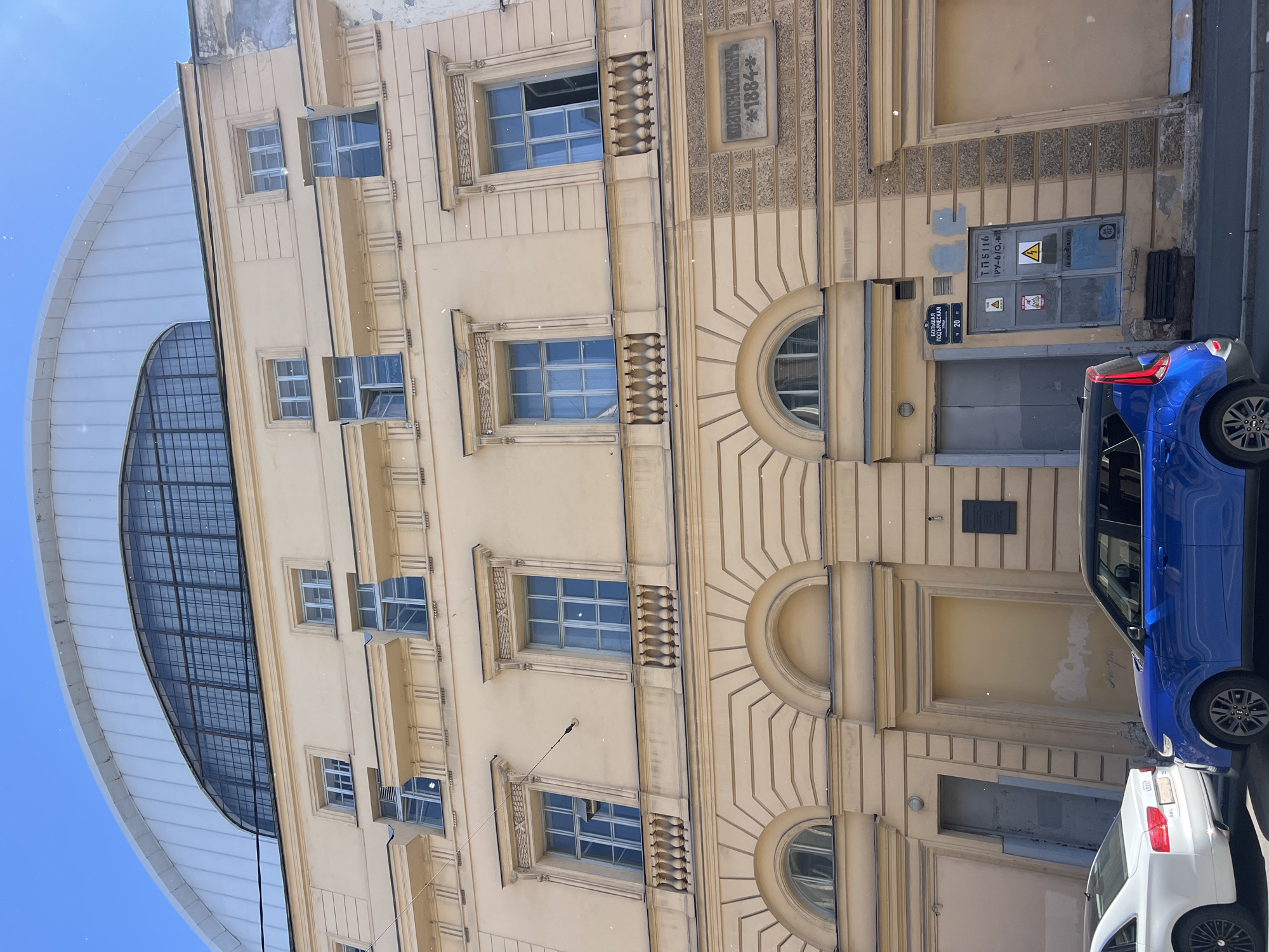 The image captures the facade of a historical building in Saint Petersburg, Russia. The building is painted in a light yellow hue and features classical architectural elements such as arched windows and decorative molding. The upper section of the building is capped by a distinctive large, curved, ribbed roof structure, partially obscuring a vast arched window with a metal grid. Multiple windows of varying sizes are visible on the facade, some of which are open. A plaque with text and a year, "1884", is affixed to the wall. In the foreground, two cars are parked on the street: a white sedan and a blue compact SUV. The scene is illuminated by daylight, with a clear blue sky above.