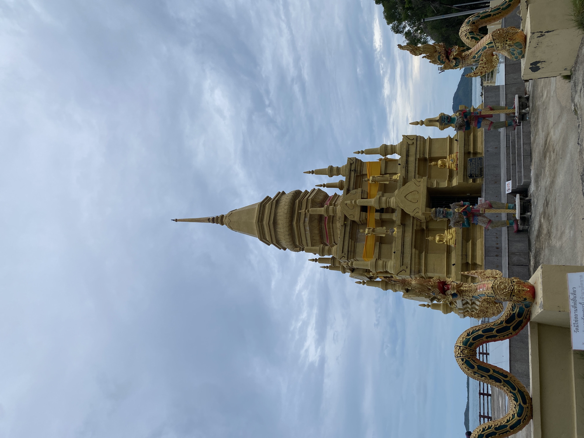 A vertical medium shot depicts a gilded stupa in Baan Thong Krut, Thailand, against a blue sky with white streaky clouds. The stupa is surrounded by ornate dragon statues. A concrete path and stairs lead up to the stupa. A sign with Thai script is visible on the right. In the background, a glimpse of the ocean and distant hills is visible. Trees are visible on the upper right of the frame.