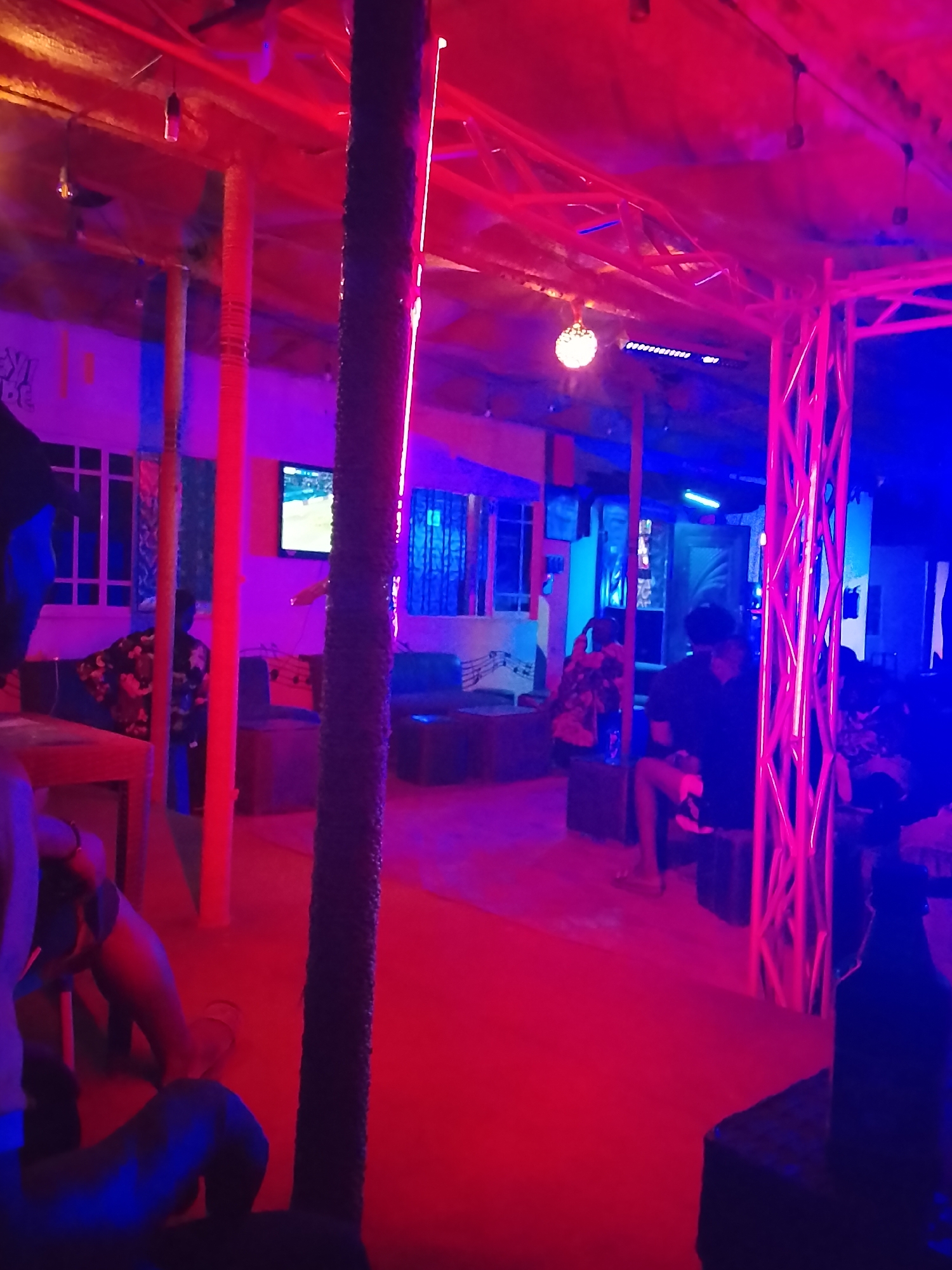 This media file depicts an indoor lounge or club environment, characterized by dynamic red and blue neon lighting that casts a vibrant glow across the scene. The setting suggests a social gathering taking place at night.
The space is supported by numerous vertical poles, some of which are wrapped in textured material, while others are painted solid red or orange. A metal truss structure, illuminated with pinkish-red neon lights, stands prominently on the right side, suggesting a stage or performance area. The ceiling appears to be constructed from planks or a similar material, with various hanging light fixtures, including a spherical, disco-ball-like light.
Several individuals are visible throughout the venue, largely seated on low couches and chairs. Their faces are mostly obscured by the lighting and angles, but their relaxed postures indicate a casual and social atmosphere. On the left side of the image, a person wearing a patterned shirt is seated, and further back, another group is observed relaxing in a blue-lit section of the room. In the immediate foreground on the left, a person is partially visible, while a dark bottle rests on a table on the right.
The floor is a bright red, reflecting the overhead lighting. Wall-mounted televisions are present, with one in the mid-ground clearly displaying what appears to be a sports event. Decorative musical notes are visible on a wall in the background, adding to the entertainment theme. On the far left wall, partial text is visible, with "VIBES" being clearly discernible, possibly part of "EASY VIBES."
The scene captures a lively, engaging social event within a venue in Yelwa, Nigeria, designed for entertainment and relaxation.