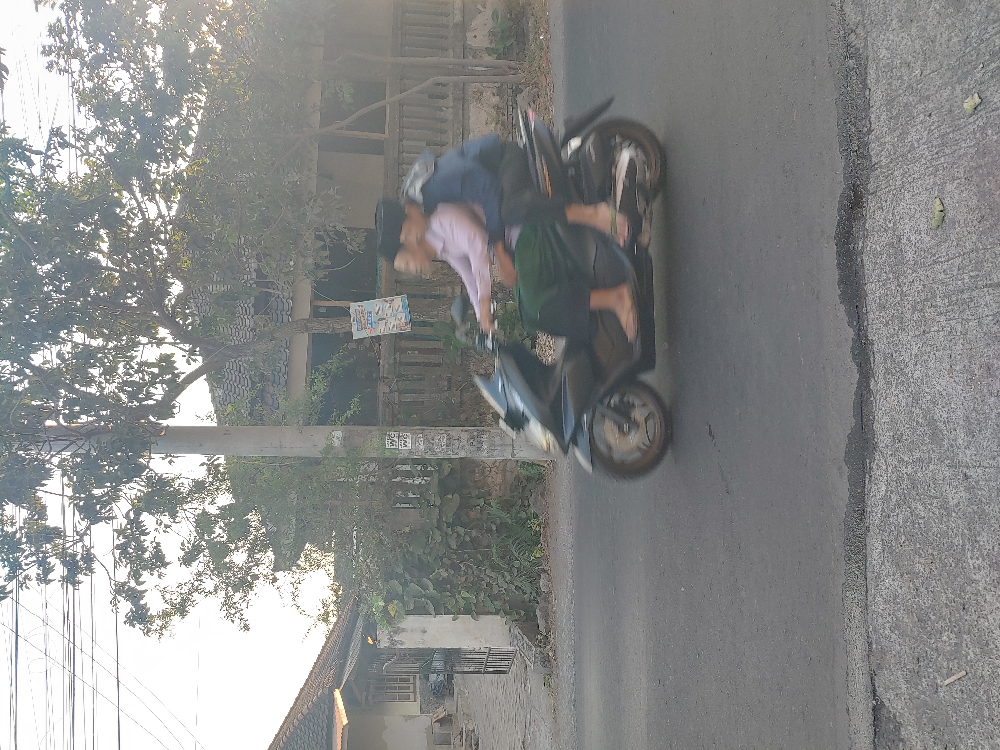 In Kembang Kerang, Indonesia, two individuals are captured in motion on a dark-colored scooter. The scooter is traveling on an asphalt road with a gray concrete curb on the left side. The person in the front is wearing a light pink long-sleeved shirt, a green sarong, and flip-flops. The person behind is wearing a dark blue jacket and black pants, with a grey scarf draped over their shoulders. Both individuals appear to be wearing black skullcaps. The background features a wall covered in green vines, a concrete utility pole with posters attached, and houses with tiled roofs. Trees with lush green foliage are visible in the mid-ground and background, and overhead, power lines crisscross the bright, slightly hazy sky. The image is characterized by motion blur, indicating movement.