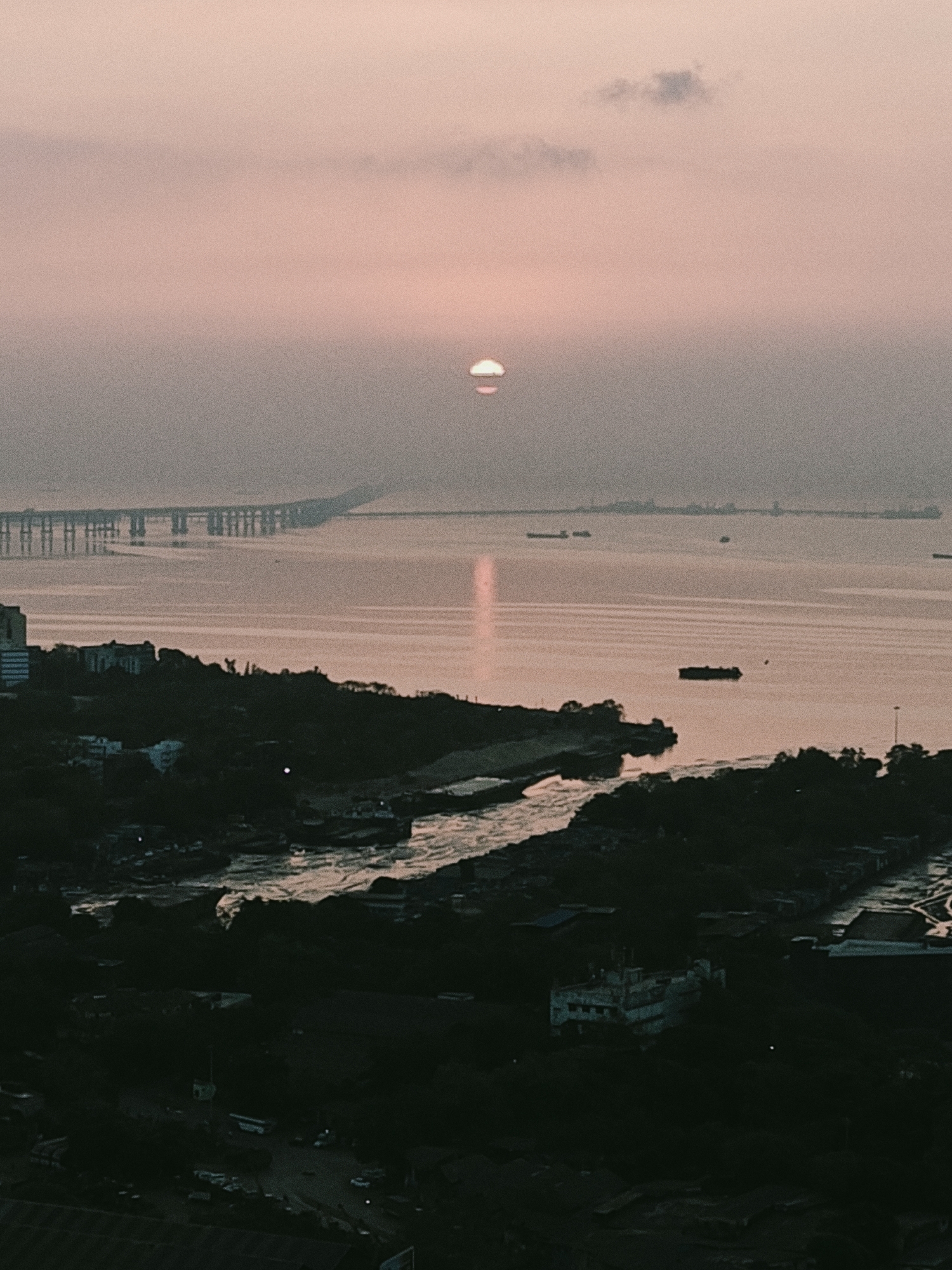 The image shows a view from an elevated position in Mumbai, India, looking out over the Arabian Sea. The sun is setting (or rising) with a pink/orange hue, partially obscured by clouds/haze. In the distance, the Bandra-Worli Sea Link bridge is visible. Several ships and boats are on the water. The foreground includes a densely populated urban area with buildings, trees, and roads. The image is somewhat dark and grainy.