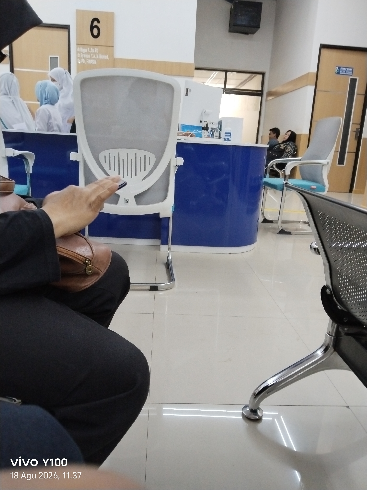 The image depicts a waiting area, likely within a medical facility, given the presence of a numbered room sign ("6") with names of medical professionals printed below. The room sign also indicates "FINASIM", potentially a medical association. In the foreground, a person is seated, wearing dark clothing and holding a brown handbag. Their right hand is visible, holding a mobile phone. Behind this person, white chairs with blue seats are arranged. Further back, a blue counter is visible, with a white dispenser-like device on it. To the right, another chair with a grey back and blue seat is prominent. In the background, two individuals are seated, one leaning against a wall. A door labeled "STAFF ONLY" is also visible on the right side of the image. The floor is tiled with a light, reflective surface. The image appears to have been taken indoors in a well-lit environment. The timestamp indicates the photo was taken on August 18, 2026, at 11:37 AM. The location is identified as Patokan, Indonesia.