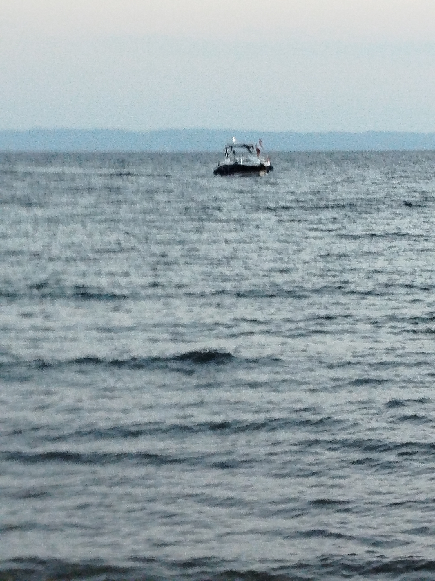 An eye-level, medium shot captures a wide expanse of choppy, grey water under an overcast sky. In the distance, a small white boat with a dark hull is visible, its silhouette sharp against the hazy blue horizon line of a landmass. The boat appears to be stationary. The water surface is textured with numerous small waves, creating a pattern of light and dark streaks. The overall impression is one of a cool, possibly early morning or late afternoon scene in a coastal area.