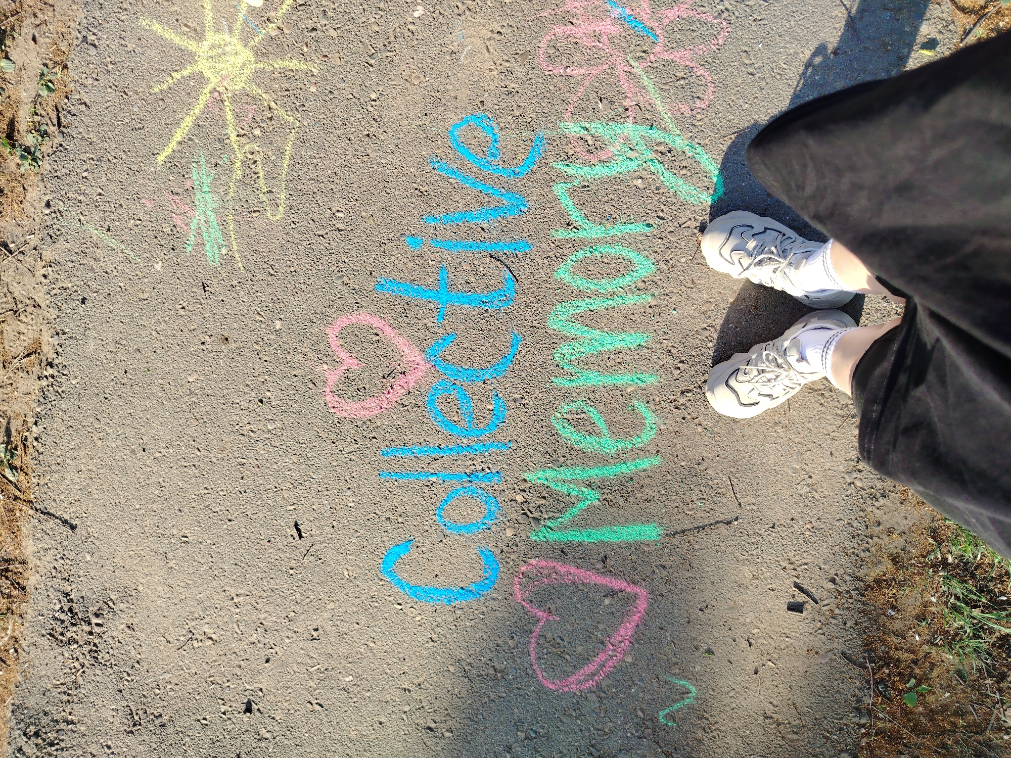 The image shows a section of paved ground, likely a sidewalk or road surface. The words "Collective Memory" are written in chalk on the ground, using blue and green chalk for the letters, and pink chalk for heart symbols. A person's feet and lower legs are visible in the upper right corner of the image. The person is wearing white sneakers and black shorts. A grassy area is visible along the right edge of the image. A yellow chalk sun is drawn on the sidewalk on the upper left. Location information is unavailable to me.