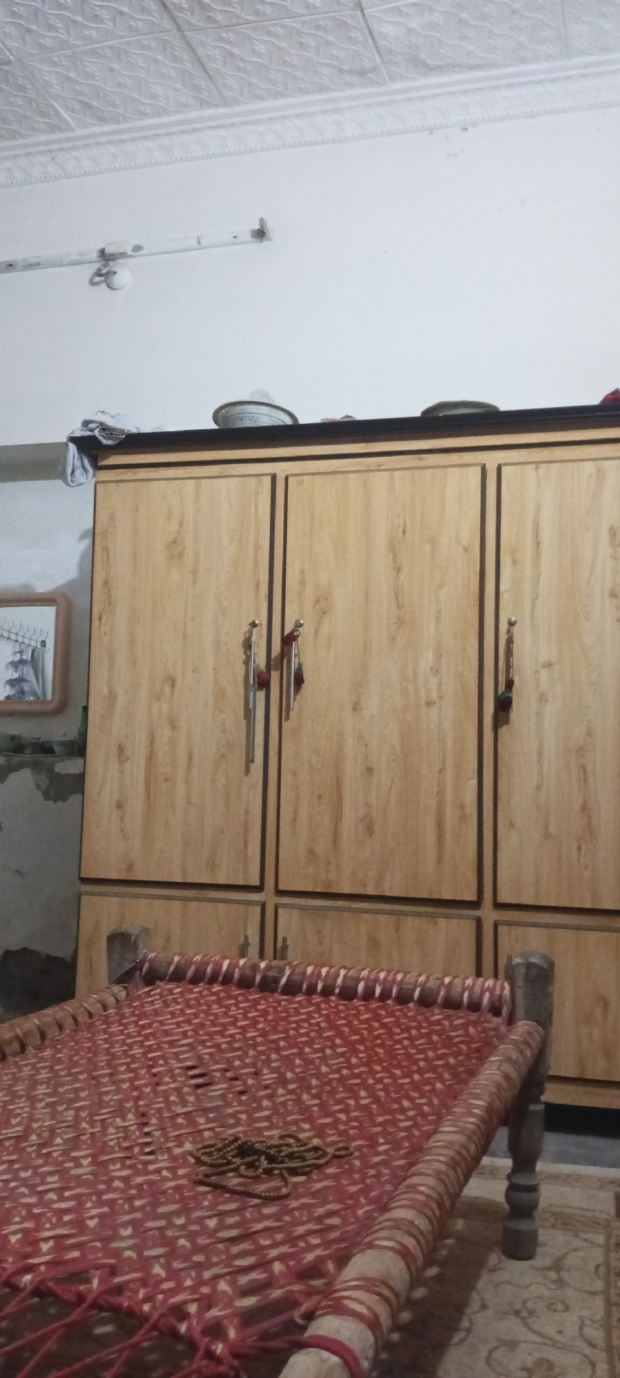 The image shows a room in Katlang Tehsil, Pakistan, featuring a large, wooden wardrobe against a wall. The wardrobe has three sections and is topped with hats and clothing items. In front of the wardrobe is a traditional woven bed (charpai) with a strand of beads on it. The room has a simple ceiling, and a framed picture hangs on the wall next to the wardrobe. The overall setting suggests a modestly furnished interior.