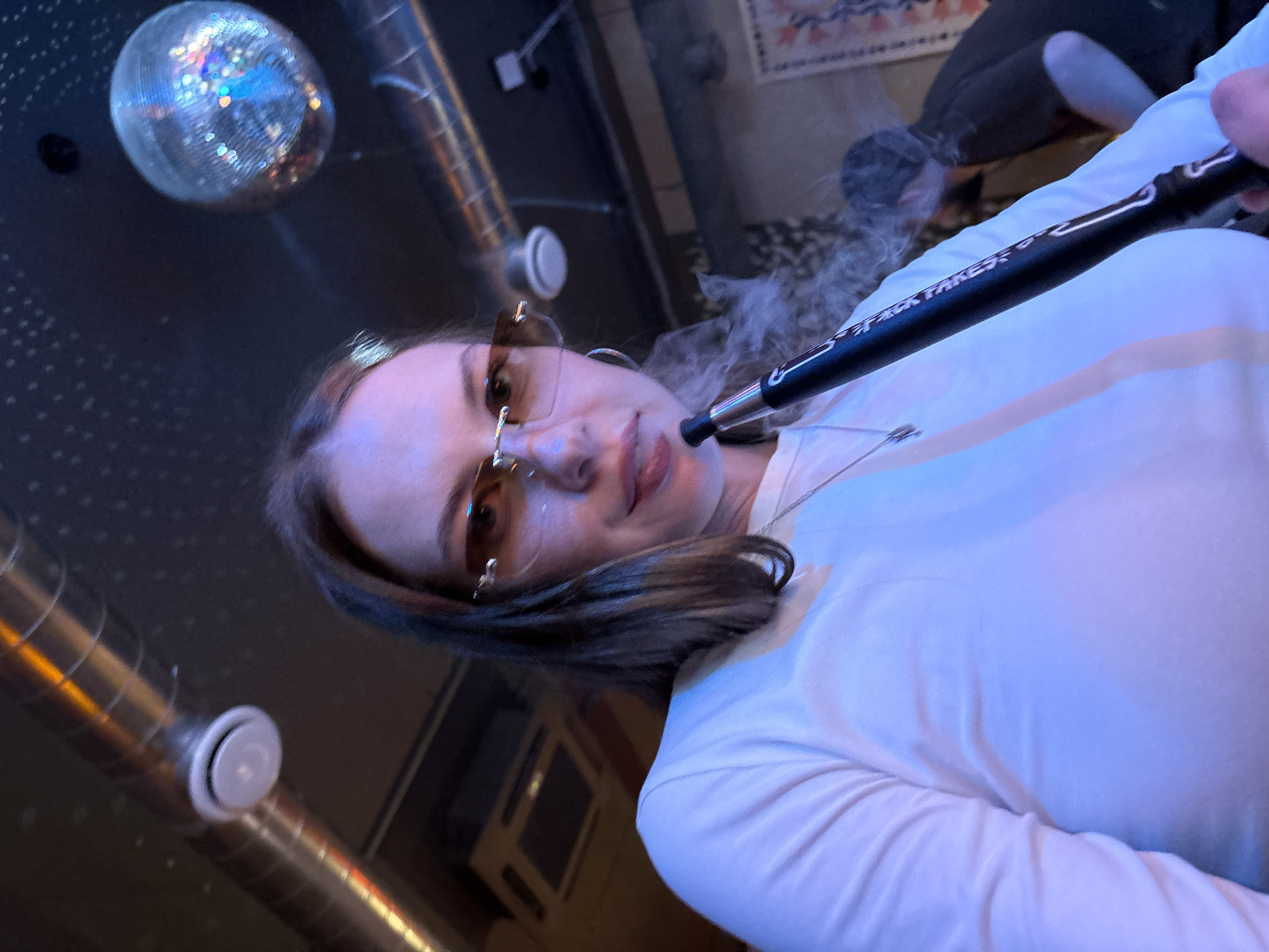The image depicts a young woman with brown hair, wearing a white long-sleeved shirt and tinted sunglasses, holding a hookah mouthpiece and exhaling smoke. She is positioned in the foreground, with her face partially obscured by the smoke. In the background, another person with dark hair is visible. The environment appears to be an indoor setting with industrial-style ventilation ducts, a disco ball hanging from the ceiling, and a patterned tapestry on the wall. The lighting is dim and colored, suggesting a nightlife atmosphere. The location is identified as Perm, Russia.
