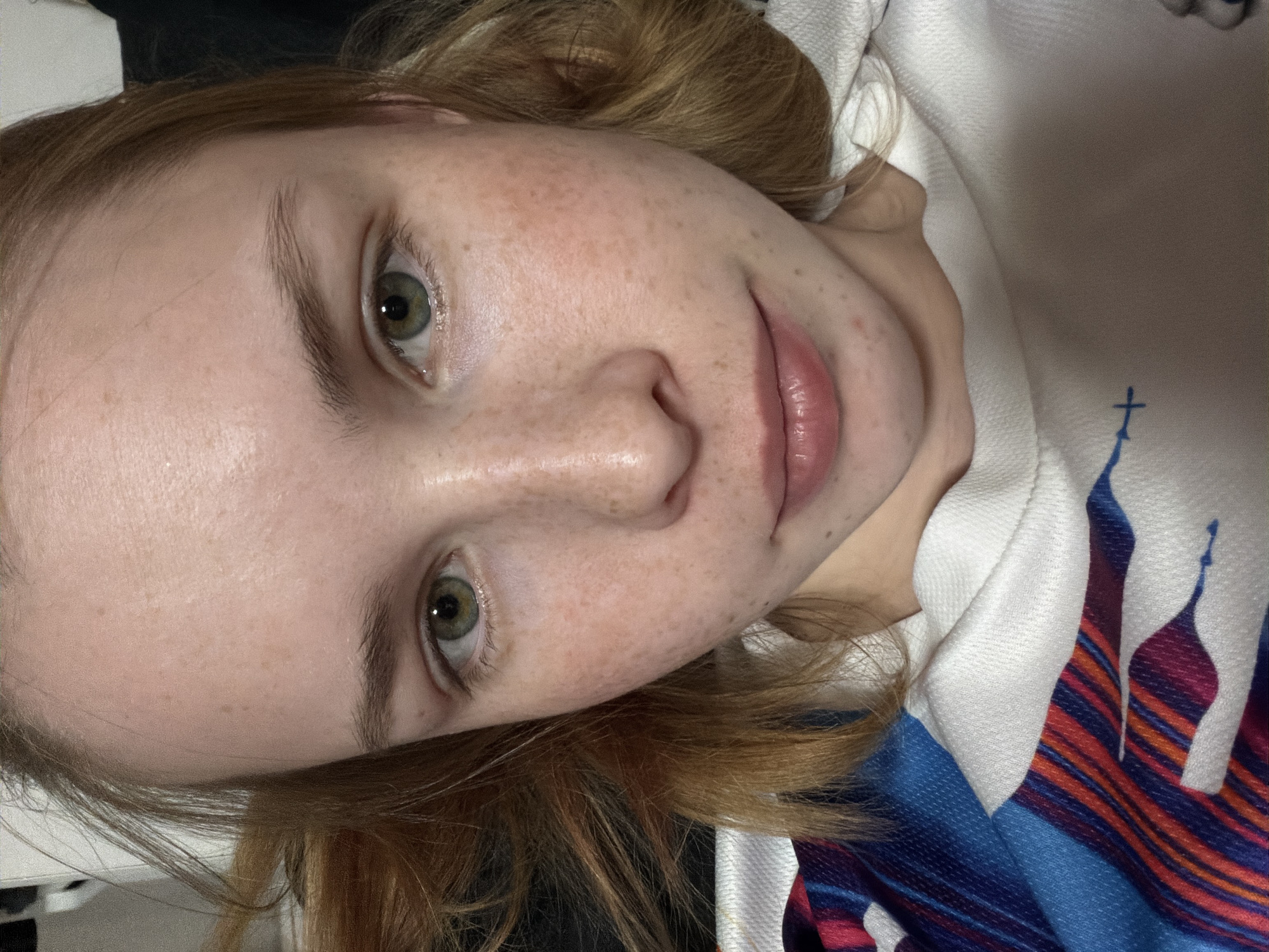 A close-up, rotated image shows a fair-skinned person with freckles and reddish-blonde hair. Their eyes are green. They have pale pink lips. The person is wearing a white top with a blue, red, and orange striped design on the fabric, which also features an image of a building with a spire and cross. The environment is not identifiable, thus cannot be determined if the location is in the city of Berezniki, Russia.