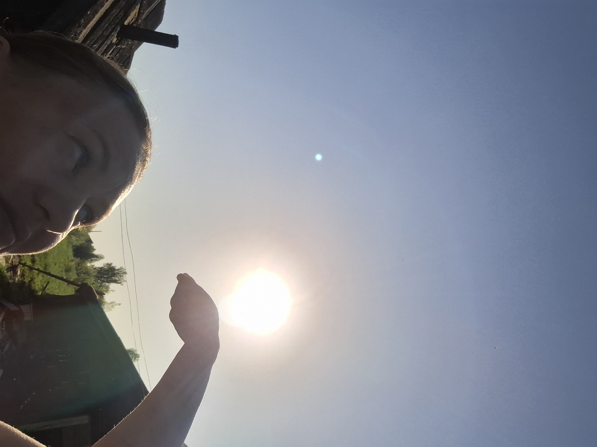 A selfie oriented in a way that suggests the photograph was taken looking upward towards the sky. A person's face is visible in the top left corner of the image. An arm is extended toward the sun, which appears as a bright white circle in the lower center of the image. The sky is a clear gradient of blue, becoming lighter towards the horizon. The lower left corner also contains a part of what appears to be a green-colored roof. The location is Шешурга, Russia.
