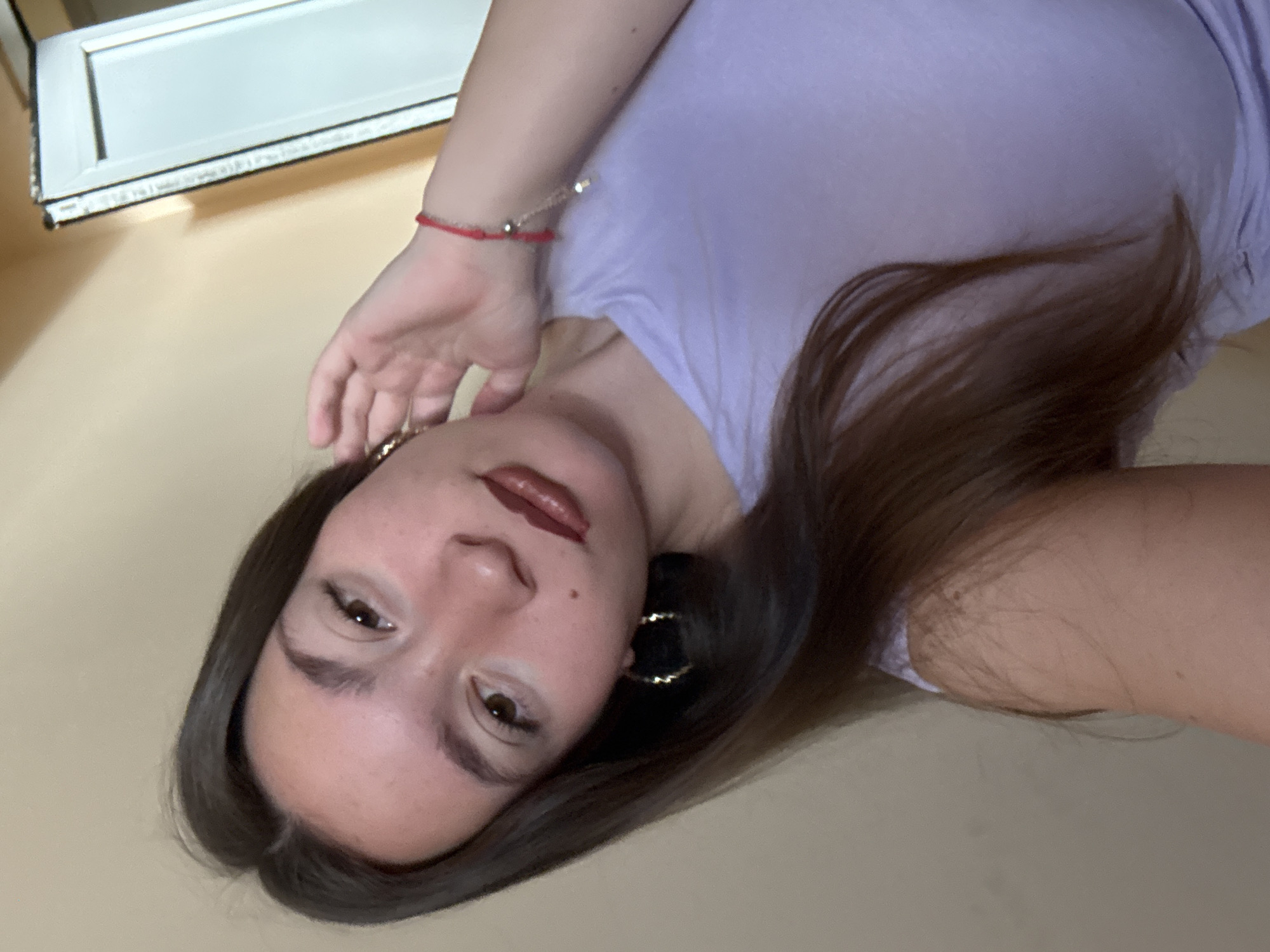 A young woman with long, dark brown hair is looking at the camera with her head tilted. She is wearing a light purple t-shirt and large hoop earrings. A red string bracelet with a silver charm is visible on her right wrist. Her makeup includes dark lipstick and mascara. The background appears to be an indoor setting with a plain, light-colored wall and a white doorframe on the right. The lighting suggests it is daytime. The specific location in Sofia, Bulgaria is not discernible from the image.