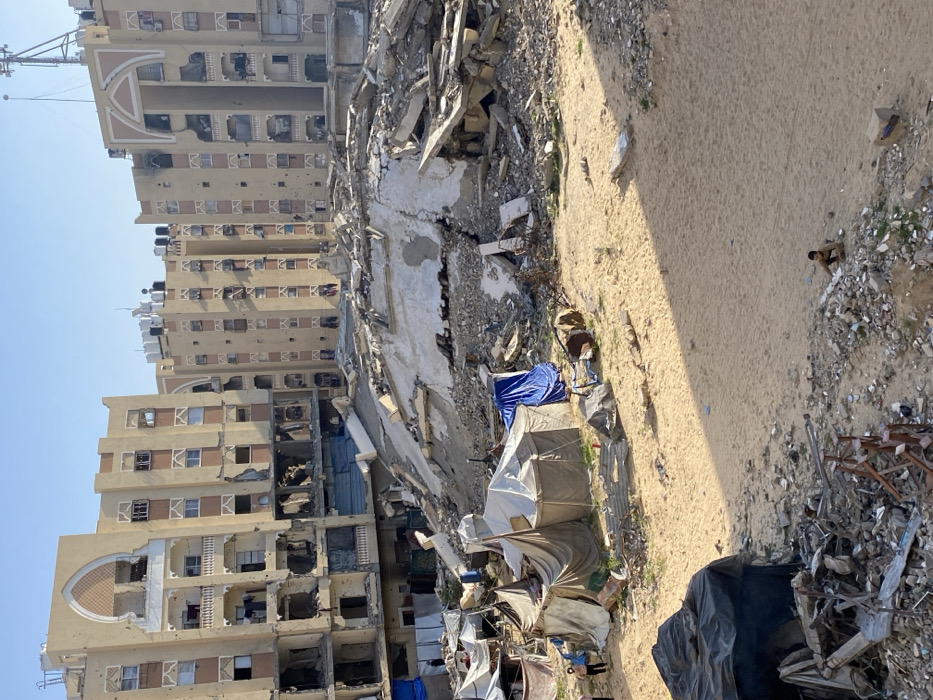 The image depicts a severely damaged urban landscape in Khan Yunis, Palestinian Territory, under a clear blue sky. In the background, multiple multi-story residential buildings, constructed from light-colored concrete or stucco, exhibit extensive damage. Their facades are riddled with numerous circular impact holes, and large sections of walls and balconies are missing, exposing internal structures. Water tanks and communication equipment are visible on their rooftops.
In the midground, a substantial mound of pulverized concrete and rebar constitutes the remains of a collapsed structure, forming a significant rubble pile. The ground in the foreground and lower midground is sandy and uneven, strewn with scattered debris, including twisted metal and broken concrete.
To the left of the central rubble, a cluster of makeshift shelters, constructed from various tarpaulins (including blue and tan) and other salvaged materials, stands on the sandy ground. Several individuals are present: two figures in light clothing are near these shelters on the left, and a small child in dark clothing is seated on a low mound of sand and debris in the bottom right corner of the frame. Long shadows stretch across the sandy ground from an unseen source to the right.