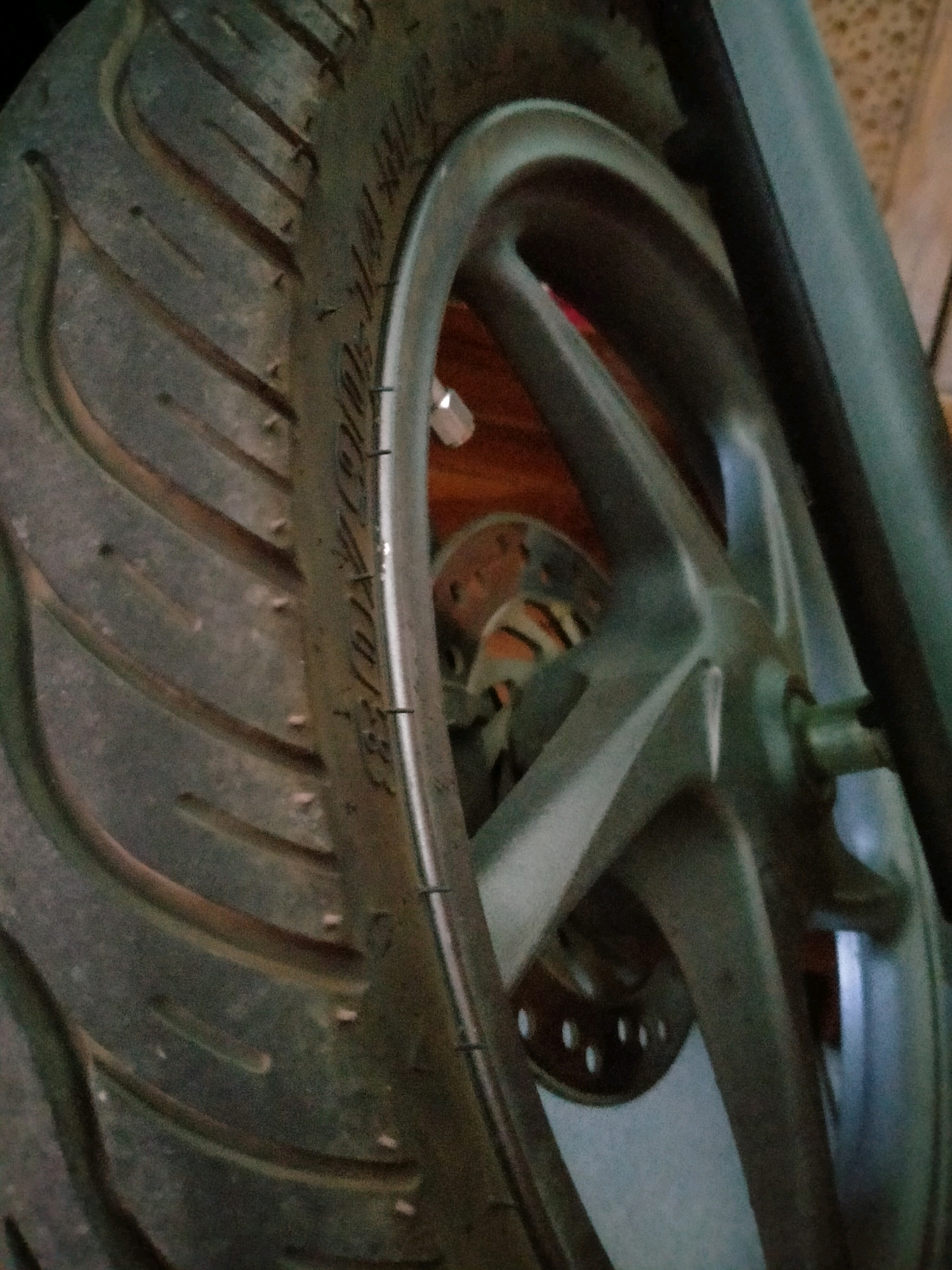 The image is a close-up, angled shot of a motorcycle wheel and tire. The tire has a prominent tread pattern with several diagonal grooves. Some small, raised dots are visible on the surface of the tire. The wheel itself appears to be made of a dark, metallic material, with spokes radiating from the center. Part of a brake disc is visible behind the spokes, with several circular holes. The background is blurred, but suggests an indoor setting with wooden textures and possibly some light-colored tiling or patterned material. The lighting is dim, casting shadows and highlighting the contours of the wheel and tire. There are no people visible in the image. The scene is a static, detailed view of a mechanical component, likely a motorcycle's front or rear wheel.