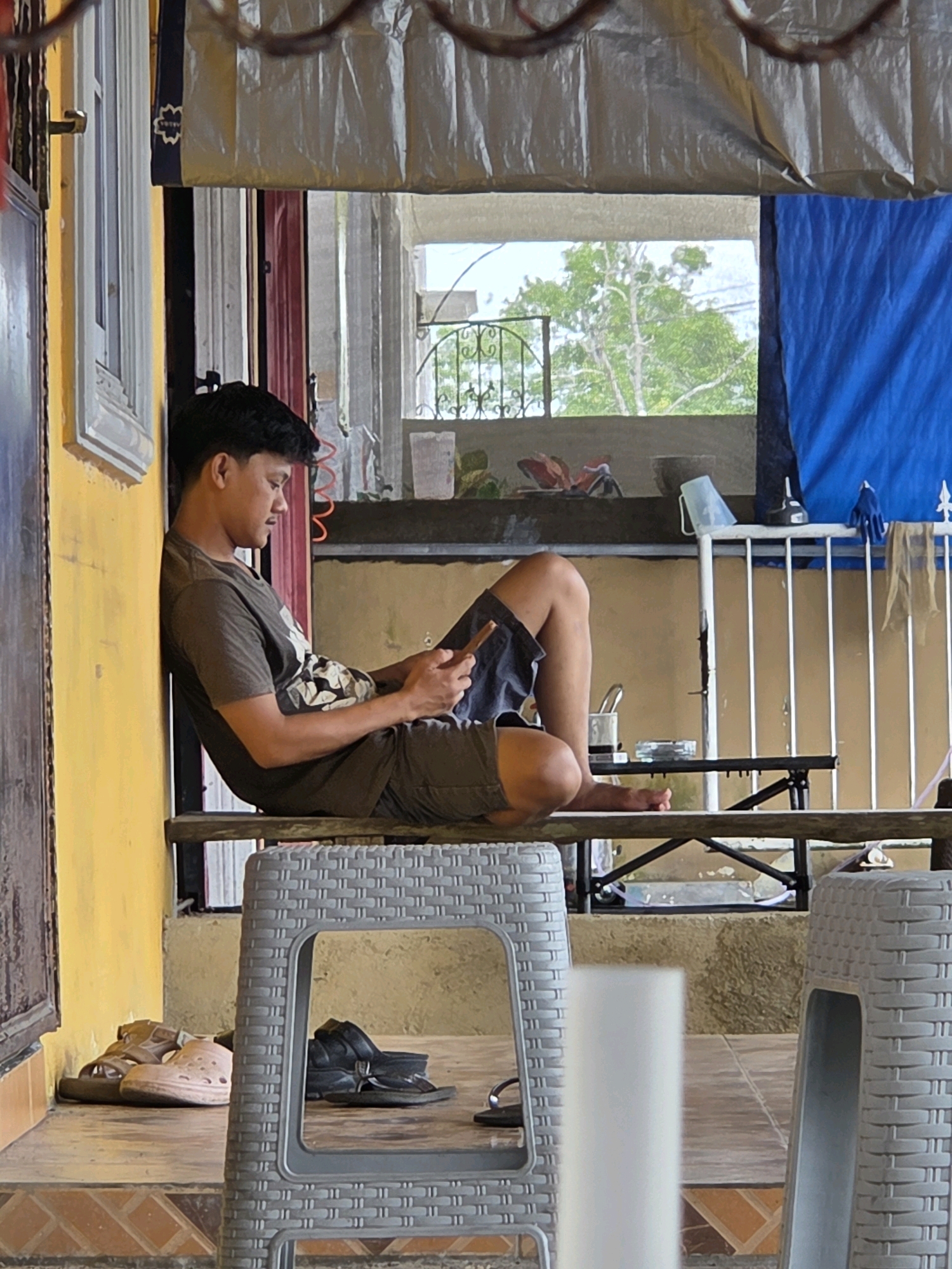 A young man is seated on a wooden bench leaning against a yellow wall, looking at his smartphone. He is wearing a dark green t-shirt with a graphic print and dark shorts. His legs are crossed, with his right leg bent and resting on his left knee. In front of him, on a lower level, is a grey plastic chair with a woven texture. To the left of the bench, on the floor, are a pair of brown sandals and a pair of beige clogs. To the right of the bench, behind the grey chair, is another grey plastic chair. In the background, through an open area with a metal railing, trees and foliage are visible. A blue tarp is draped on the right side of the background. The scene appears to be an outdoor seating area, possibly a cafe or restaurant, in Tanjung Pinang, Indonesia. The lighting suggests it is daytime.