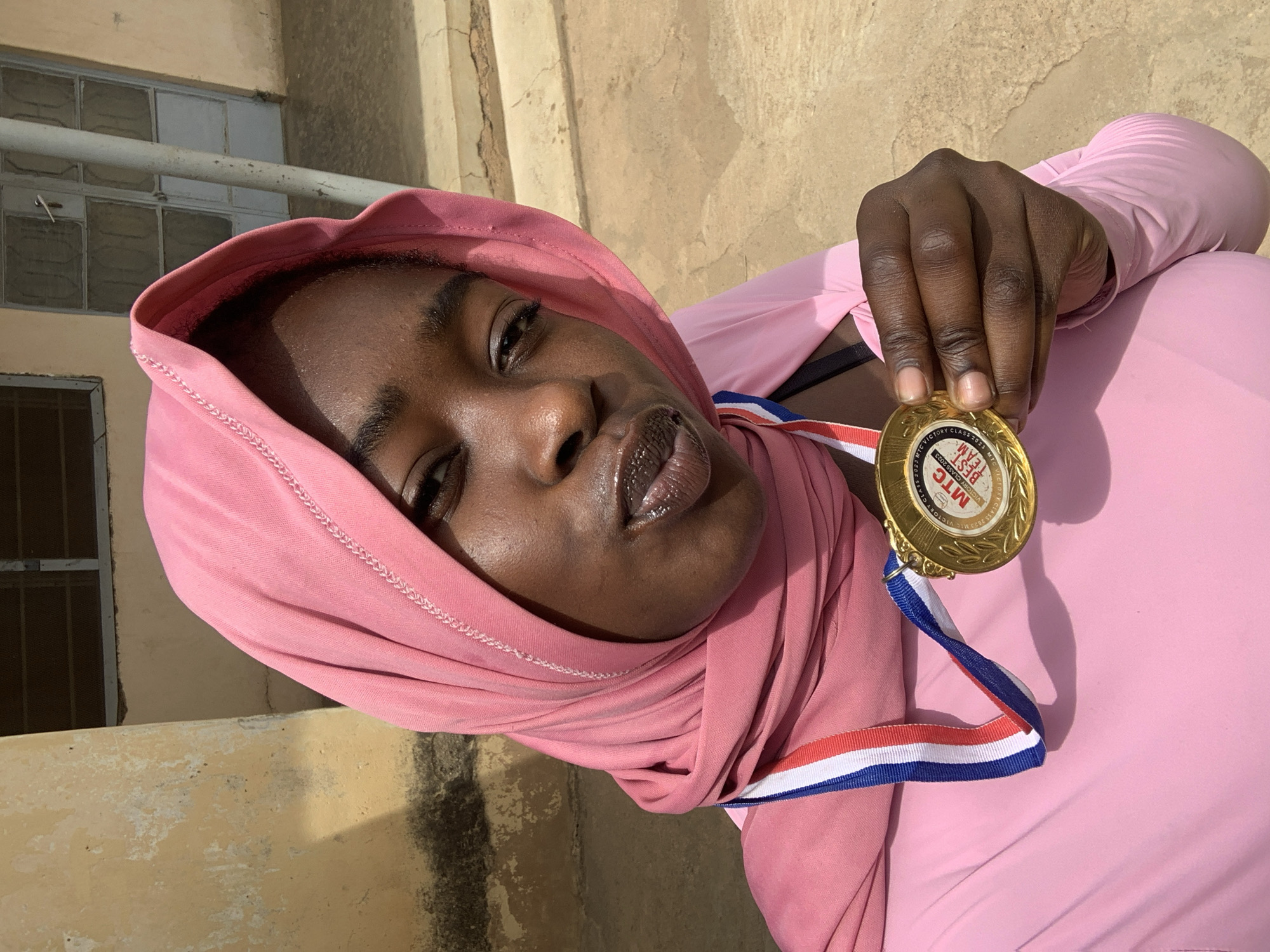 A young woman wearing a pink headscarf and a matching long-sleeved shirt is holding a gold medal with a red, white, and blue ribbon. The medal appears to commemorate "MIC VICTORY CLASS 2023". The woman's skin is dark, and her lips are glossy. She is looking at the camera. The background features a beige or light-yellow wall and a window with metal bars. The scene seems to be outdoors with natural light. The location is identified as Rafin Par, Nigeria.