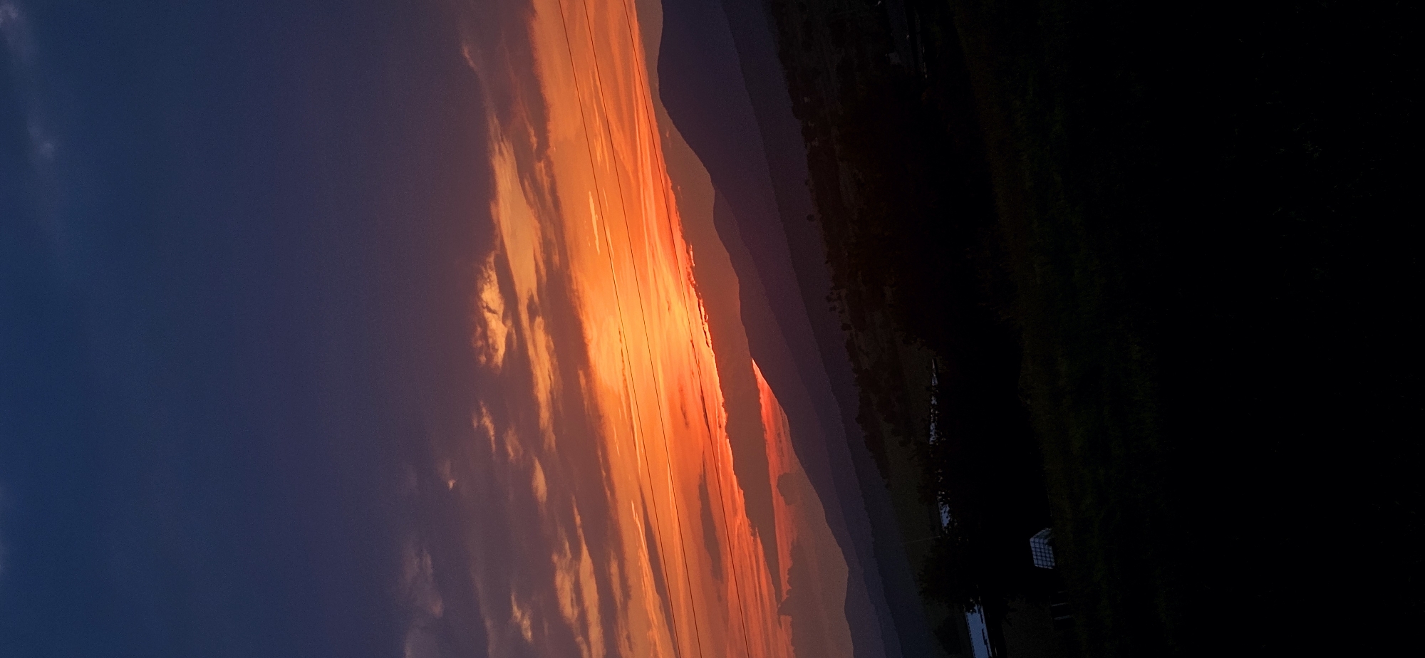 The image presents a wide landscape taken during sunset in Momin Prohod, Bulgaria. The sky occupies the upper two-thirds of the frame, displaying a gradient from deep navy blue at the zenith, transitioning to purple, dark red, and then to a vibrant band of orange and bright red along the horizon. Horizontal cloud formations are distributed across the sky, appearing dark against the brightest orange light or catching the red and orange illumination. Multiple thin, dark horizontal lines, consistent with power lines, traverse the mid-section of the sky. Below the sky, several layers of mountains are silhouetted, appearing as dark, undulating masses against the luminous horizon. The midground consists of dark, indistinct terrain with scattered trees and vegetation. The foreground is heavily shadowed, primarily showing dense, dark foliage. Faint architectural elements, including a light-colored, gridded structure, are subtly visible within the dark midground on the left side. No individuals or animals are present.