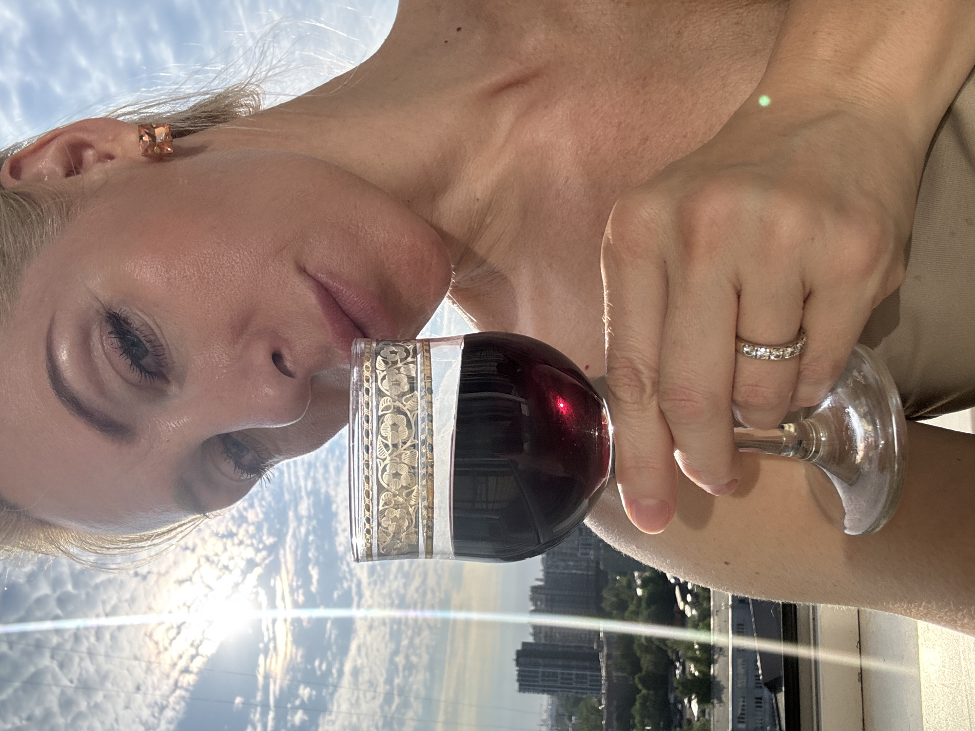 The image shows a close-up of a woman holding a glass of red wine. She has blonde hair and is wearing a beige top, square orange earrings, and a diamond ring. Her right hand is holding the stem of the wine glass. The background appears to be a city skyline seen from a high vantage point, with buildings and trees visible. The sun is shining brightly, creating lens flare and illuminating the clouds. The woman's face is partially obscured by the wine glass. The scene suggests a relaxed or celebratory moment, possibly on a balcony or rooftop overlooking the city. The location is identified as Unknown, Russia.
