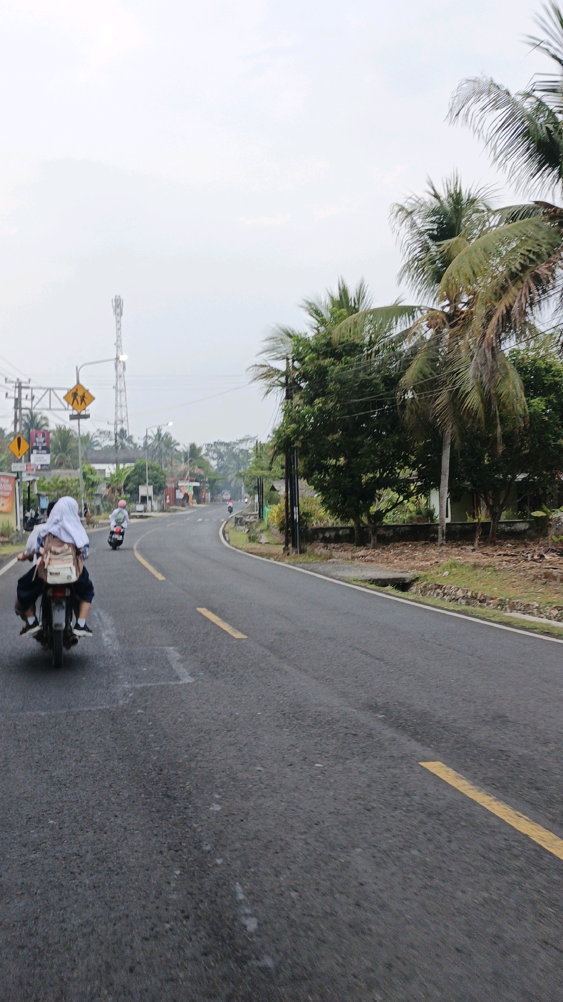 The image depicts a road scene in Sukaresik, Indonesia, with a focus on two individuals riding a motorcycle on a paved road. The rider and passenger, likely students given their attire, are seen from behind, traveling away from the camera. The passenger wears a white hijab and a backpack. The motorcycle is dark in color. The road curves to the left and is marked with yellow lines. On the left side of the road, there are traffic signs, including a pedestrian crossing sign. A cell tower is visible in the distance, along with palm trees and other foliage. The sky is overcast. The overall environment suggests a rural or suburban setting.