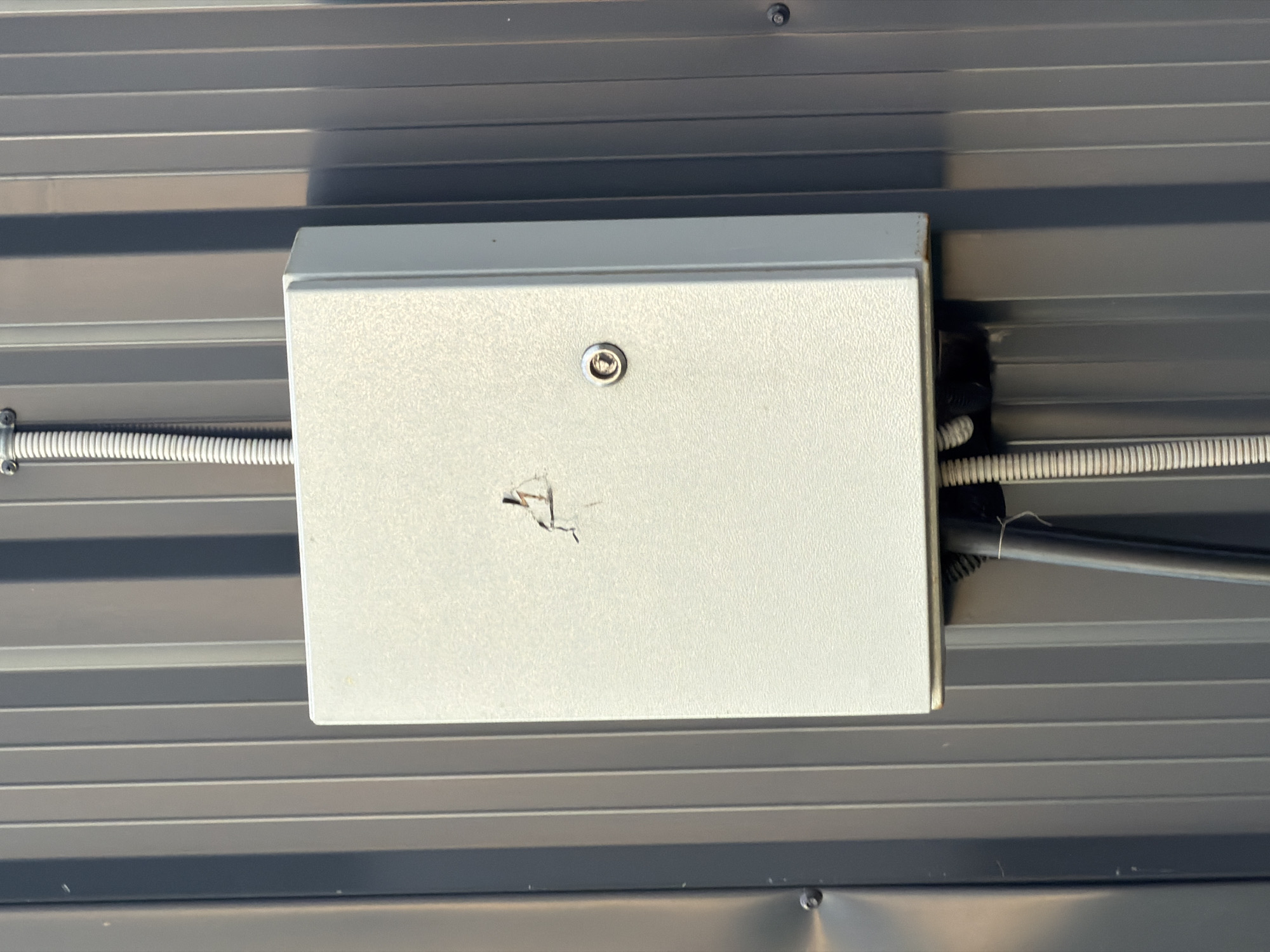 A white electrical box is mounted on a ribbed, dark grey metal wall. The box has a keyhole and a small damaged area on its front surface. A ribbed conduit runs from the top of the box and is attached to the wall, and two black cables emerge from the bottom of the box, also connected to conduit. The scene is outdoors. The location is identified as Bol'shaya Ven'ya, Russia.