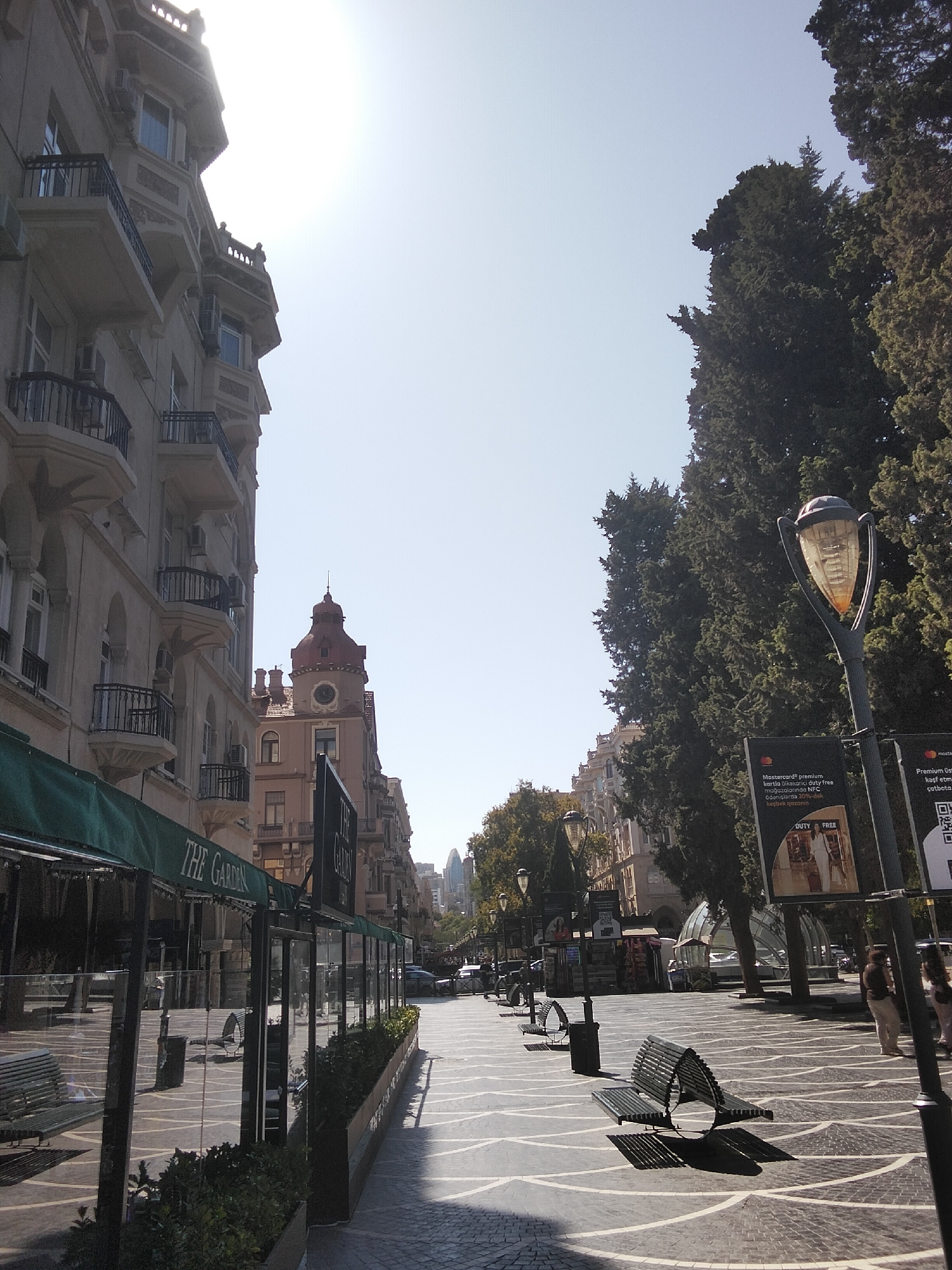 The image depicts a street scene in Baku, Azerbaijan, likely in the city center given the architecture and the presence of pedestrian walkways. On the left, a tall, ornate building with balconies and arched windows dominates the foreground, its facade illuminated by bright sunlight. To the right of this building, a clock tower with a red roof stands out.
The street itself features a distinctive paving pattern and is lined with trees on the right side. Several lampposts are visible, some with decorative glass shades. Various benches are placed along the walkway. In the middle ground, a cafe or restaurant with green awnings and glass enclosures is visible. Further down the street, the cityscape continues with more buildings and trees.
The sky is clear and bright, suggesting a sunny day. Two individuals are partially visible on the right side of the image, walking away from the camera. One appears to be wearing light-colored pants and a dark top, while the other is in similar attire. Several signs with text are also present, including one that appears to advertise "DUTY FREE" services. The overall impression is of a well-maintained urban environment on a clear day.