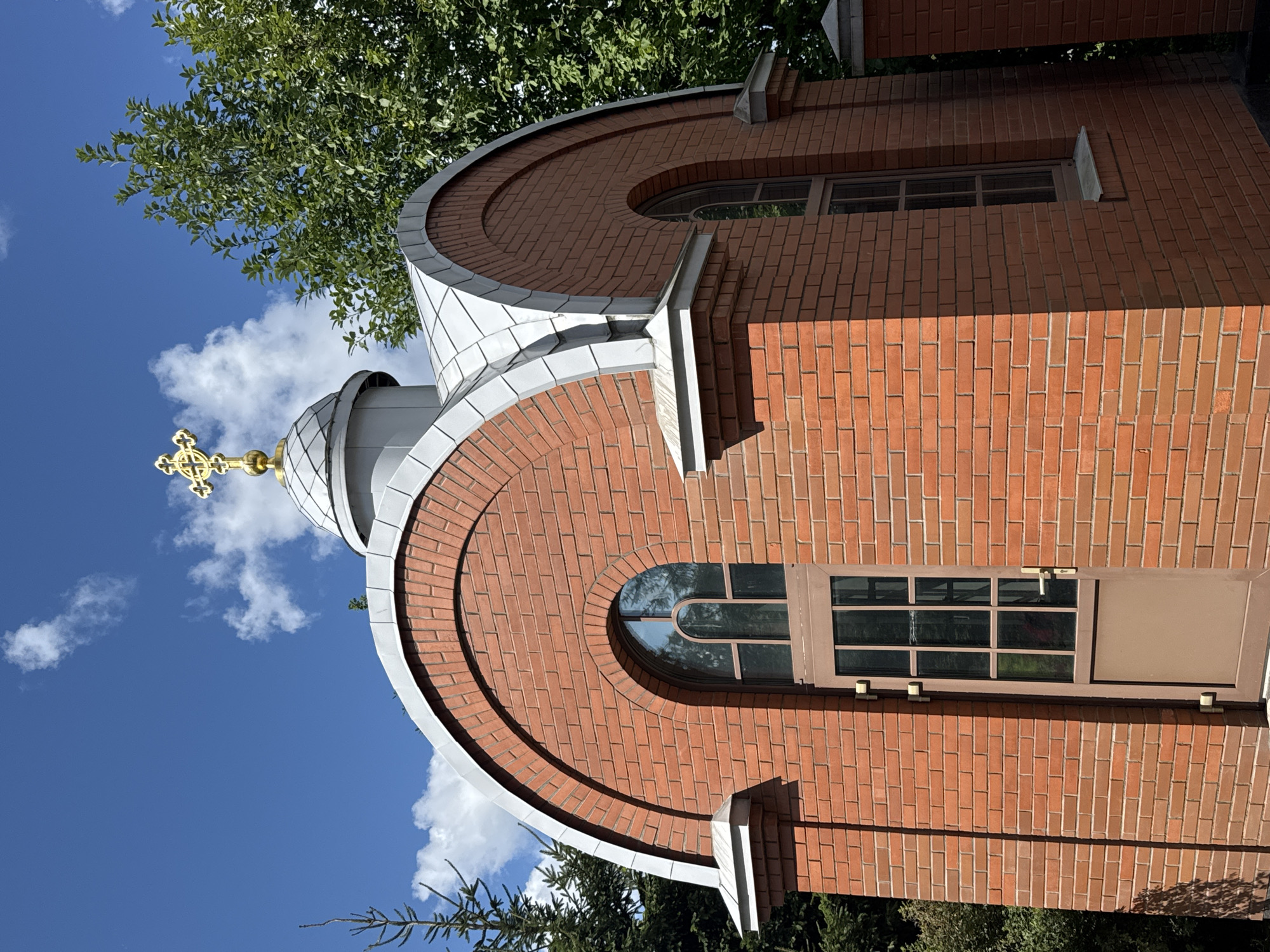The image shows a small, modern brick chapel with a silver domed roof topped by a golden Orthodox cross. The chapel has arched windows and a door with glass panes. It is situated outdoors, with lush green trees visible in the background under a bright blue sky with scattered white clouds. There are no people or animals present. The chapel appears to be well-maintained and is likely a place of worship or a memorial. Without further context or identifying markers, the specific location within Saint Petersburg, Russia, cannot be determined from the image alone.