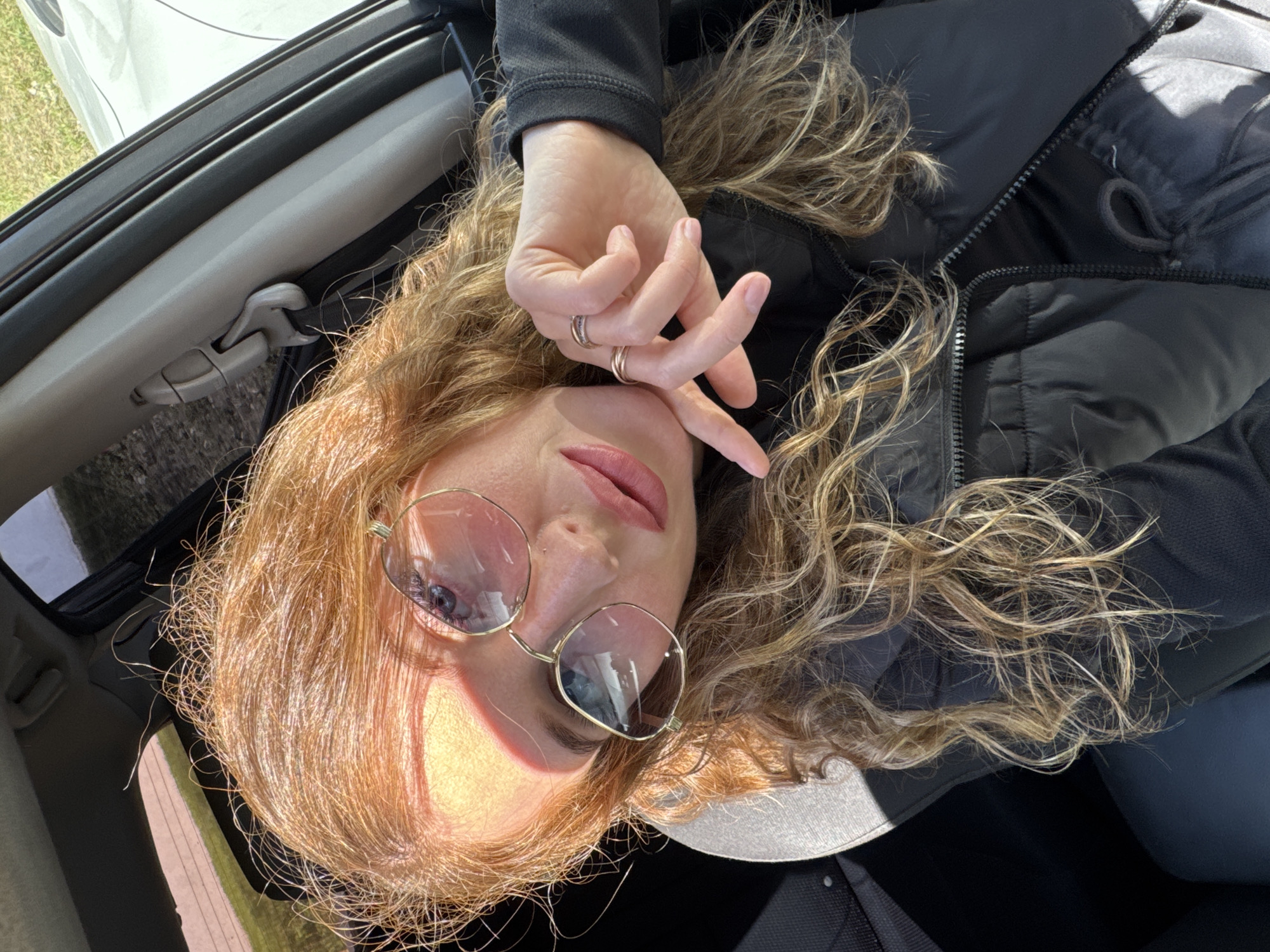 A woman is lying in the passenger seat of a car. She is wearing sunglasses and a black jacket. Her hair is blonde and curly. Her hand is raised near her face. The background outside the car is blurred, but appears to be a grassy area. The location is unknown and not Chaikovsky, Russia.