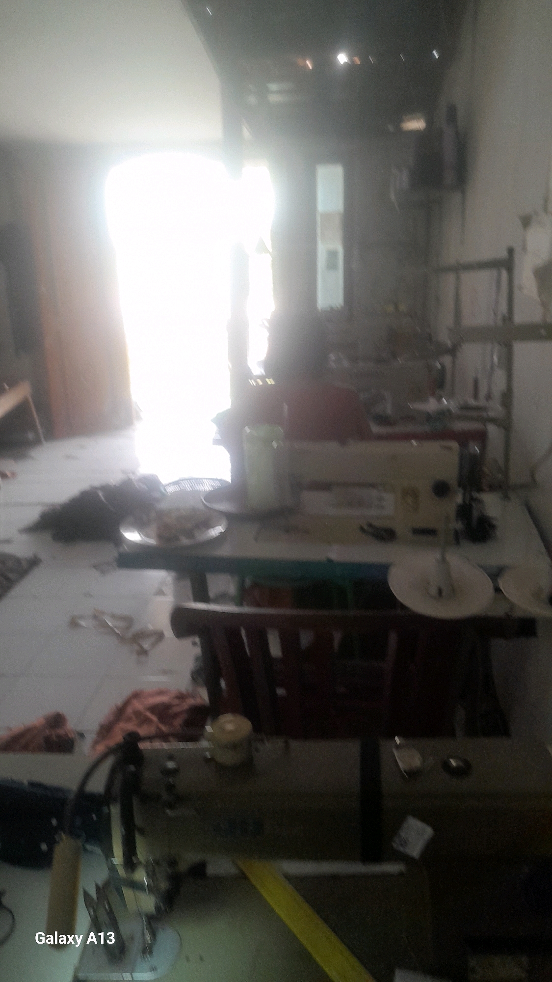 The image depicts an indoor scene, likely a workshop or tailoring shop, located in Ngelo, Indonesia. The environment is cluttered with sewing machines, fabric, and various other items related to garment production. A bright light source, possibly a doorway or window, illuminates the background, creating a strong contrast with the foreground.
In the foreground, a sewing machine is visible, with spools of thread and other sewing accessories. A dark-colored object, possibly a cat or a pile of fabric, lies on the floor near a plate of food. In the mid-ground, another sewing machine is placed on a table, accompanied by a green container and a chair. The background shows a dimly lit area with shelves and more sewing equipment.
The overall impression is one of a working space, with evidence of ongoing activity. The presence of multiple sewing machines suggests a commercial or production setting. The lighting conditions and the slightly blurry quality of the image contribute to a sense of candidness and authenticity.