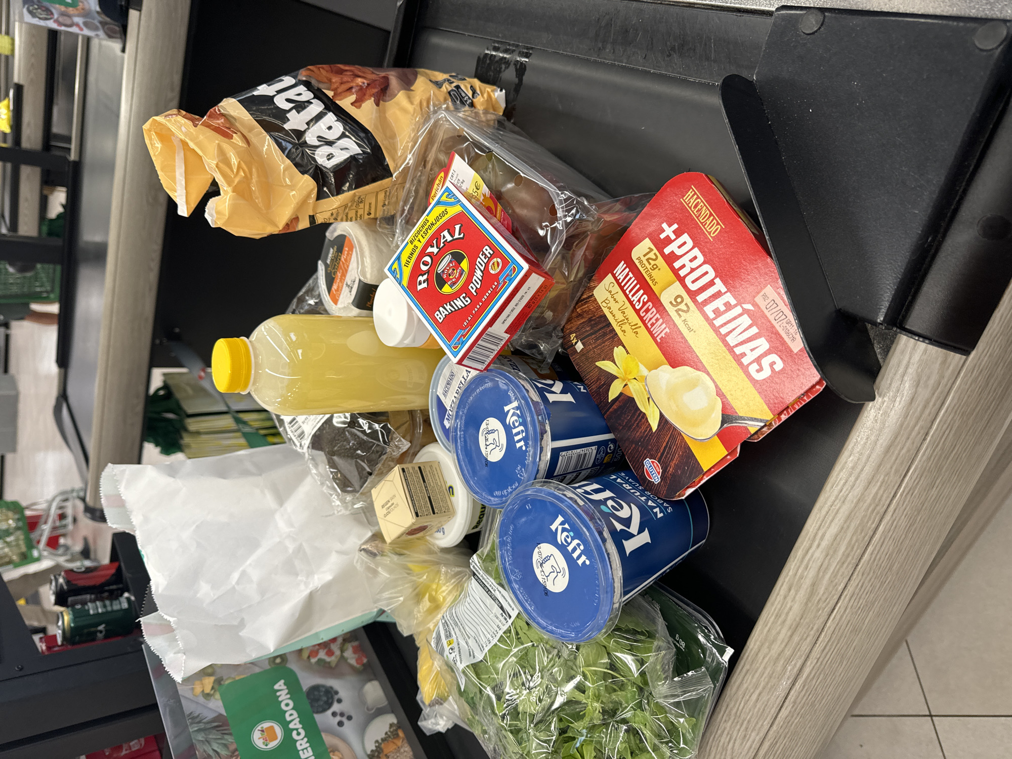 The image shows a close-up view of groceries on a supermarket checkout conveyor belt. The items include two tubs of Kefir, a bottle of yellow liquid, a bag of potato chips, a package of grapes, a box of Royal Baking Powder, a bag of leafy greens, and two containers labeled "+PROTEINAS". The background is blurred but appears to be a supermarket interior with other shoppers and shelves visible. The scene is well-lit and the objects are in focus. The presence of Spanish brand names suggests the location is in Spain. The checkout conveyor belt is made of light-colored wood.