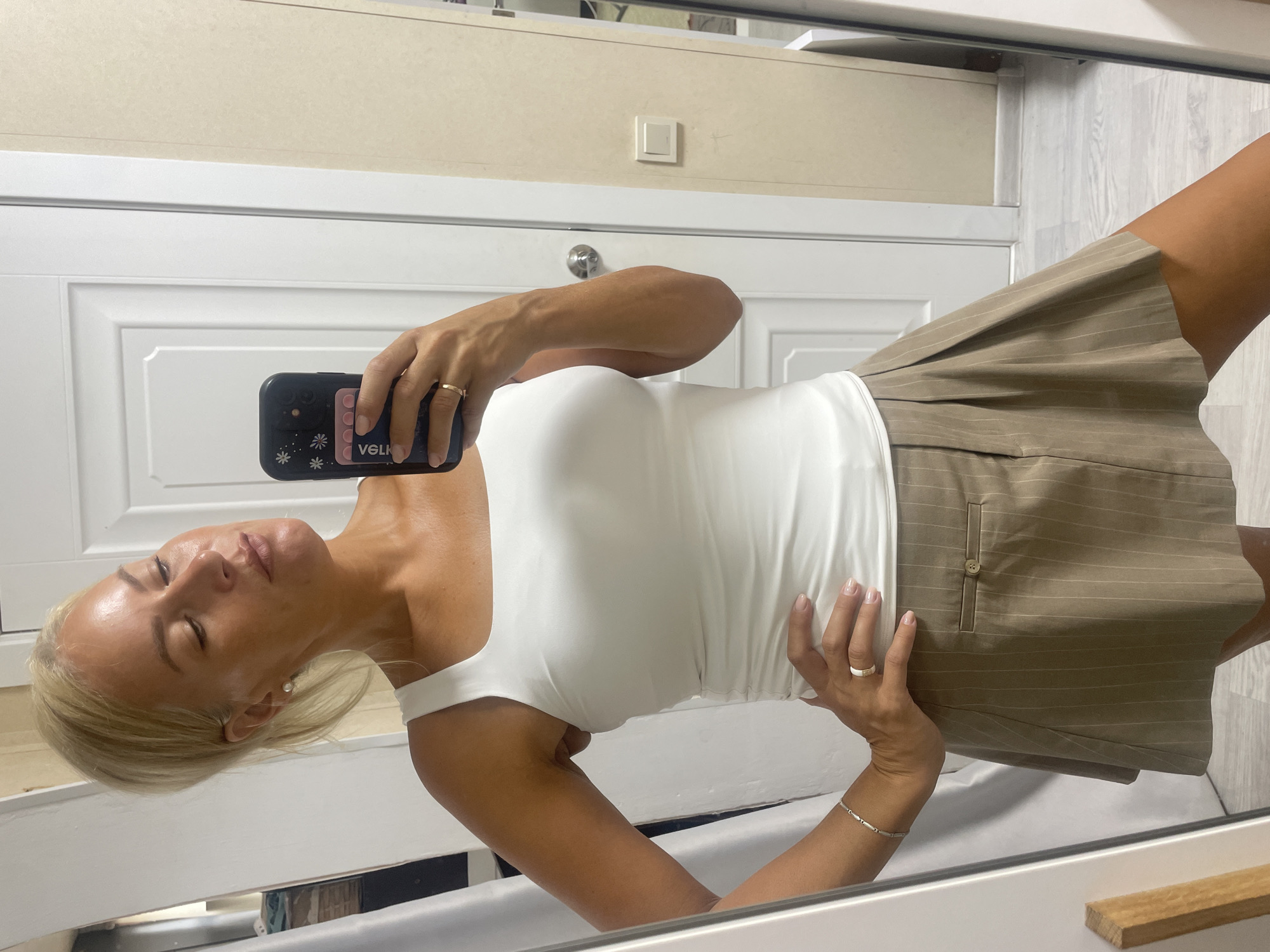 A woman with blonde hair pulled back in a ponytail is standing in front of a mirror, taking a selfie. She is wearing a white tank top with a square neckline and a tan skirt with thin vertical pinstripes. She is also wearing a silver bracelet and two rings, one on each hand. Her right hand is on her hip, and her left hand is holding a smartphone with a black case and a pink card holder. The background appears to be an indoor setting, possibly a room in a house, with a white door and a light-colored wall. There is also a glimpse of a mirror on the left side of the frame, which reflects the woman and a portion of the room. The overall impression is of a casual, self-taken photograph.