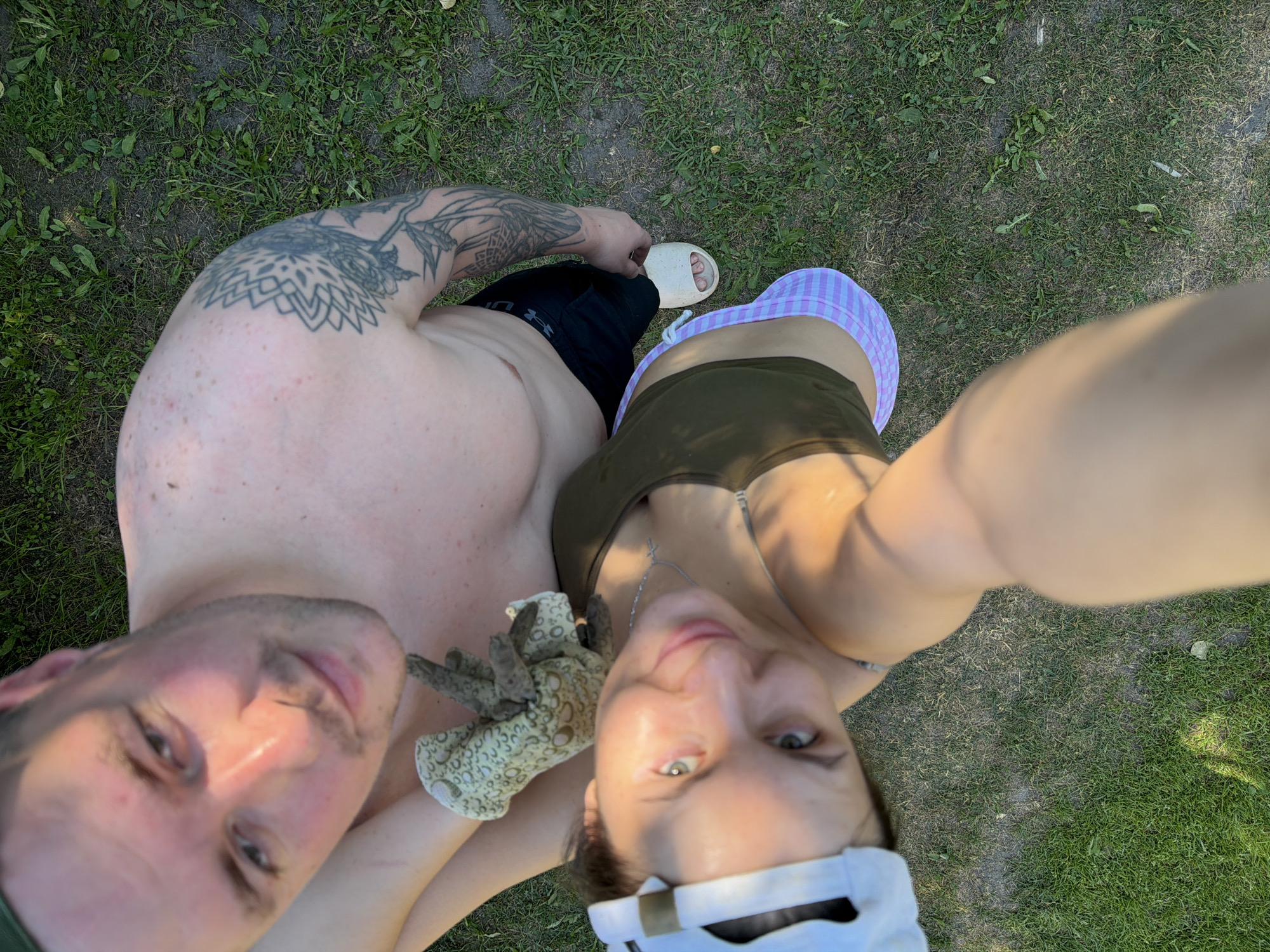 In this overhead, outdoor photograph, a couple stands on a grassy area, likely in the city of Krasny Mayak, Russia. The man, shirtless and wearing black shorts, has a tattoo on his left shoulder and upper arm. He is looking up at the camera. Beside him, a woman wearing a white baseball cap, a green tank top, and a pink and white checkered skirt is also looking up. She holds a patterned glove. The lighting suggests it is daytime, with sunlight casting shadows on the grass.