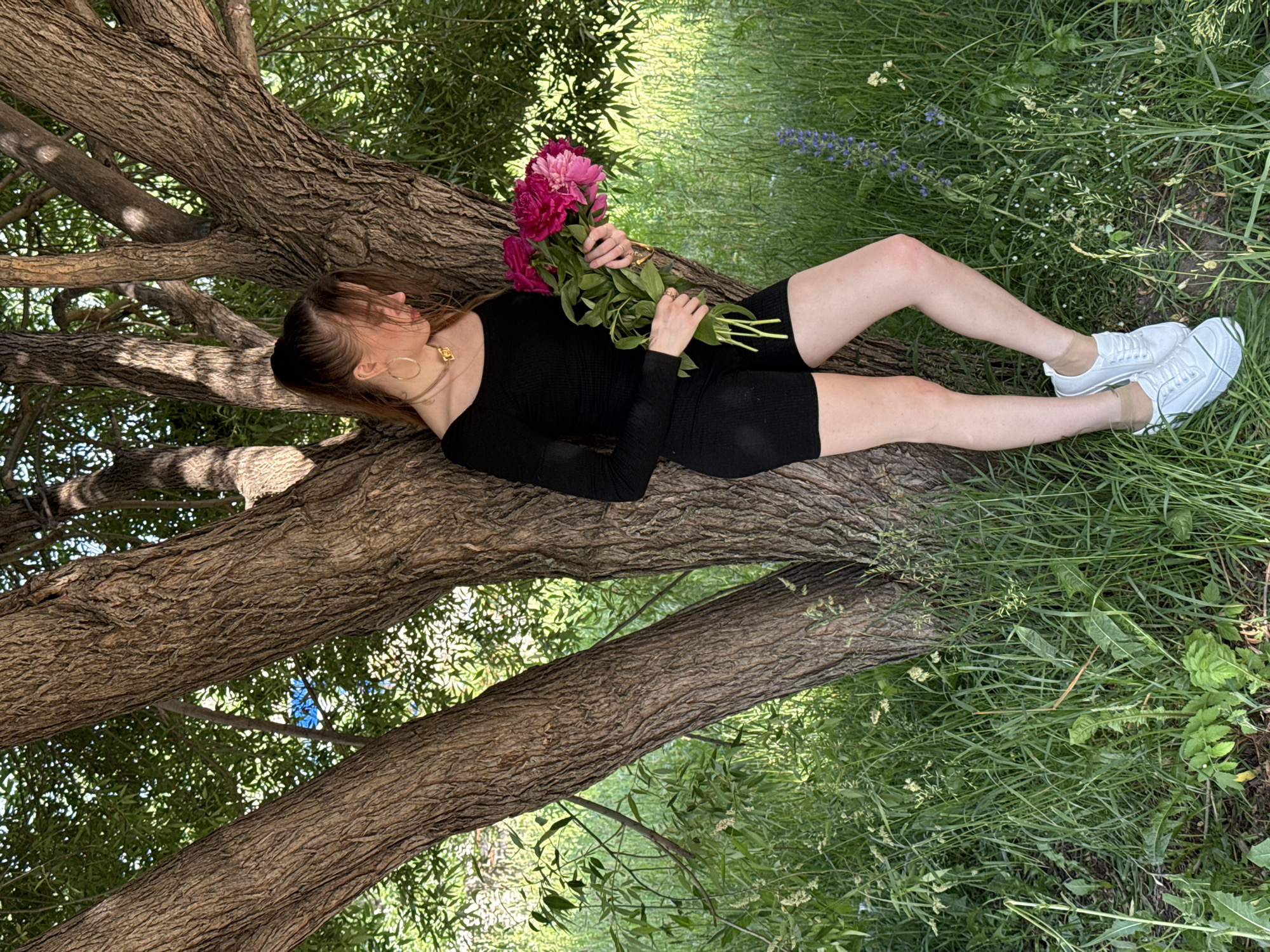 A young woman stands leaning against a large tree in a grassy outdoor area. She is wearing a black long-sleeved top and black shorts, white sneakers with tan socks. She has her hair pulled back in a ponytail and is holding a bouquet of pink and red peonies. The woman is looking down and to her right. The background is filled with green grass, trees, and foliage. The lighting suggests it is daytime, possibly late afternoon, with dappled sunlight filtering through the leaves.