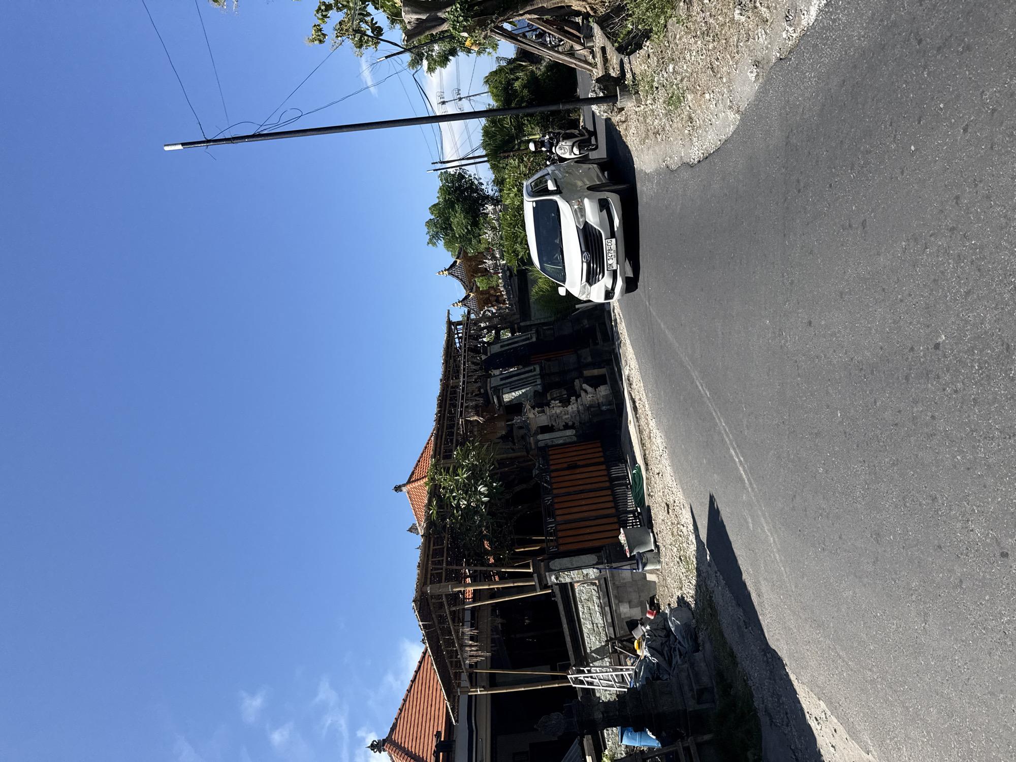 The image shows a street in Batununggal, Indonesia, on a sunny day. A white car is traveling on the road. A Balinese-style building with a partially constructed roof is adjacent to the road. A power pole is on the opposite side of the street.